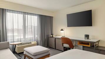 A hotel room with a bed, a desk, a chair, a small table, and a large window with curtains.