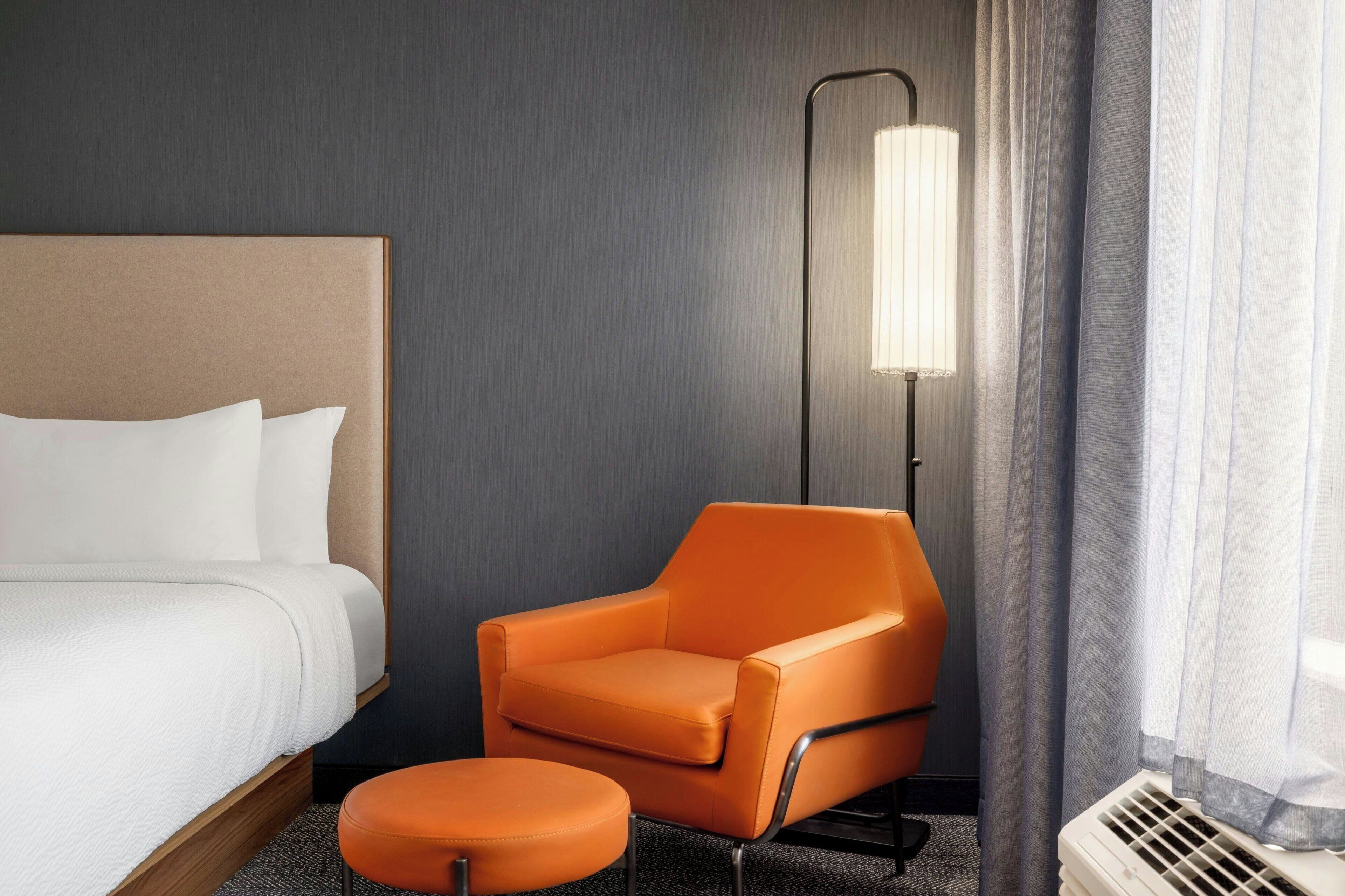 A hotel room with a bed, an orange armchair, a matching ottoman, a floor lamp, and a window with curtains.