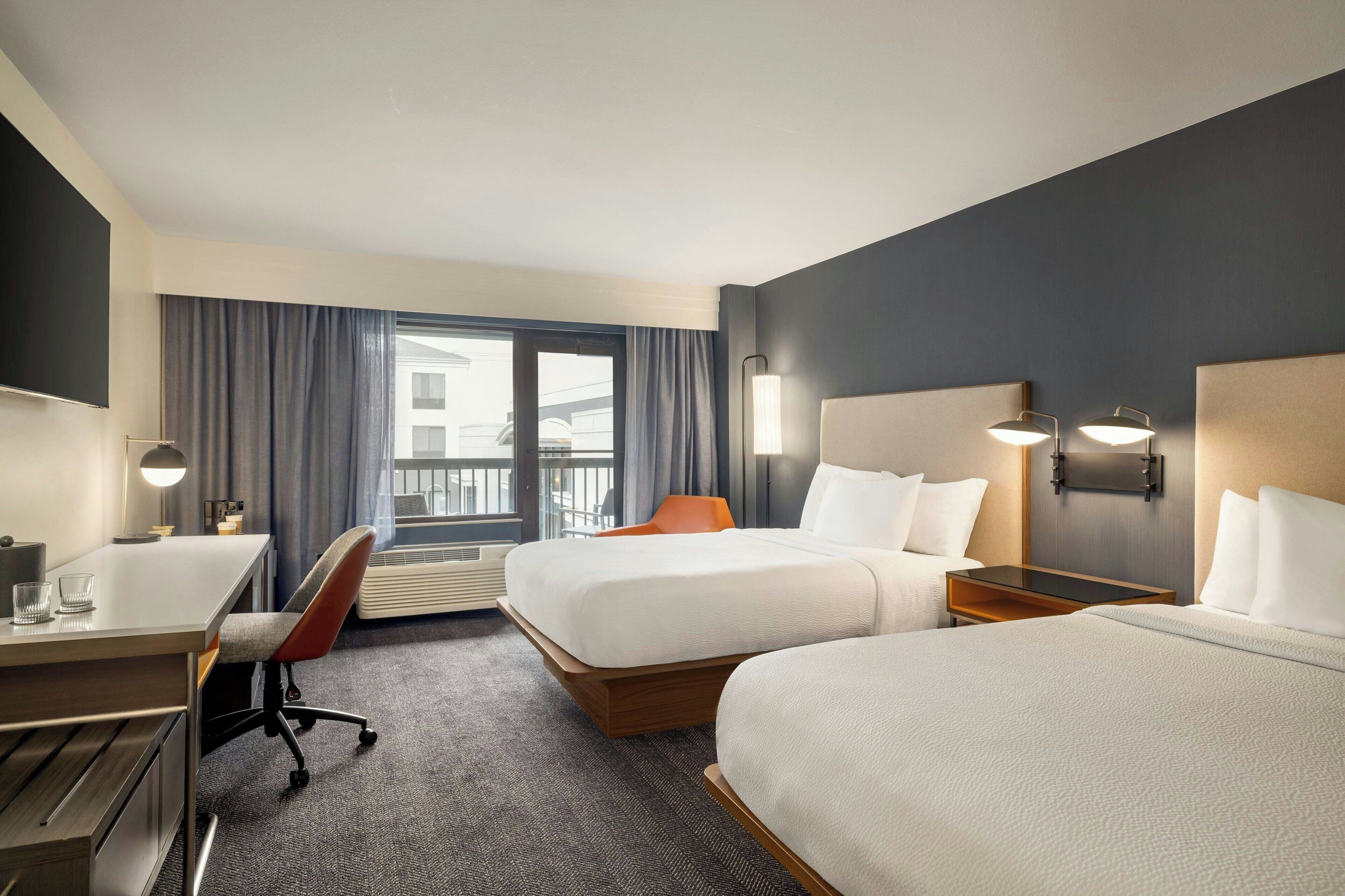 A hotel room with two beds, a desk, a chair, and a large window with curtains.