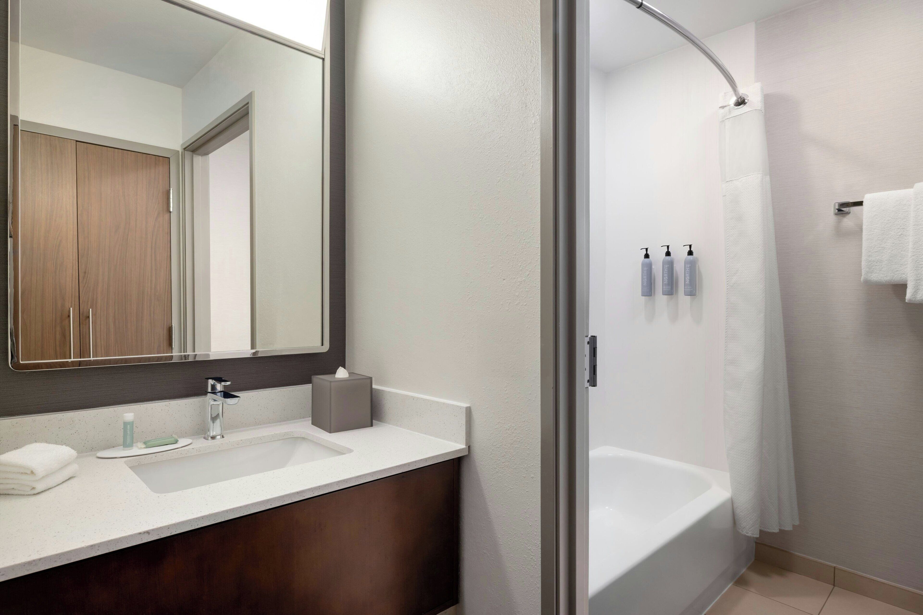 A modern bathroom with a double sink, a large mirror, a shower with a glass door, and toiletries on the wall.