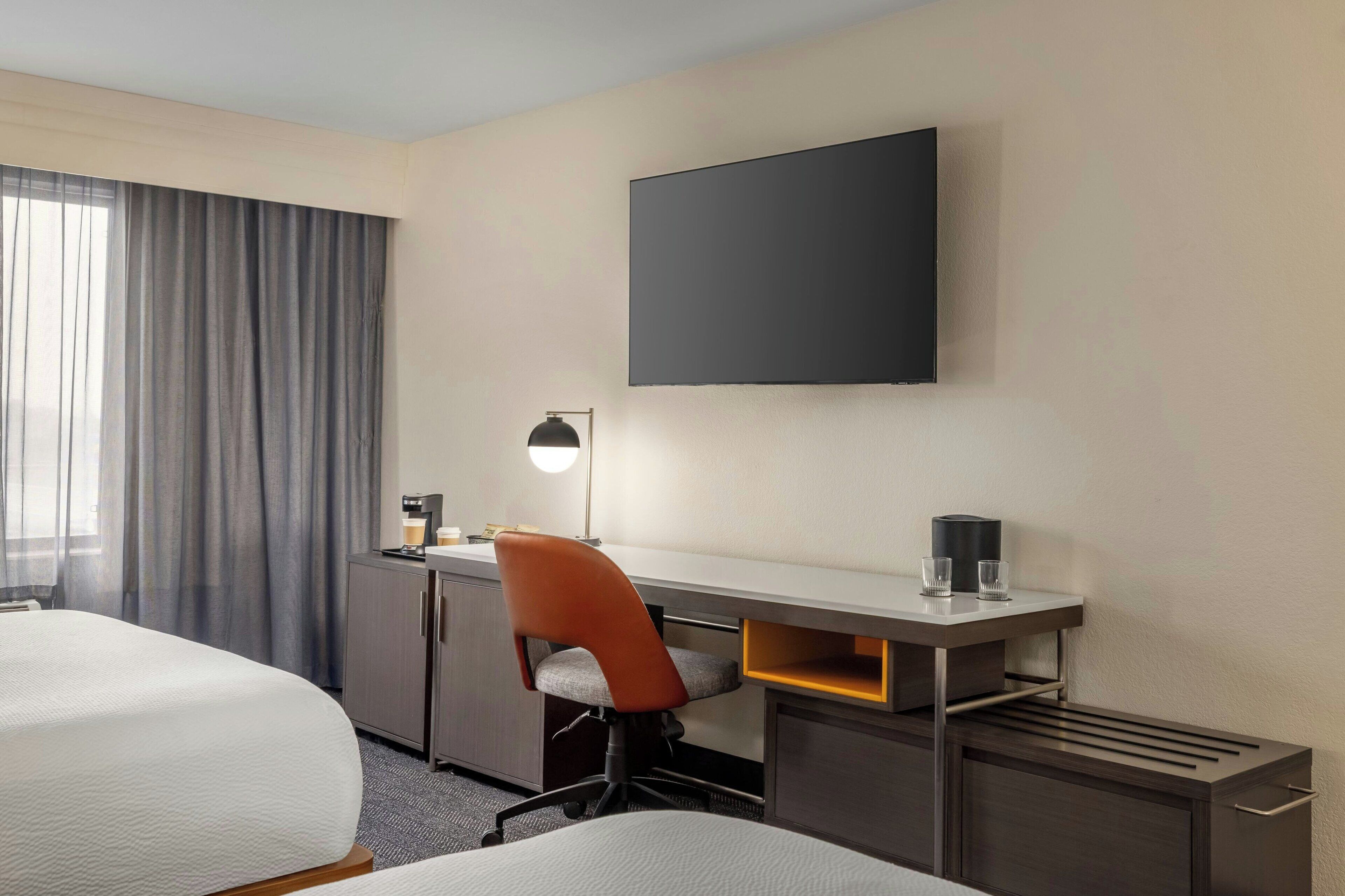 Habitación de hotel con dos camas, un escritorio con silla, televisor de pantalla plana, lámpara y cafetera.