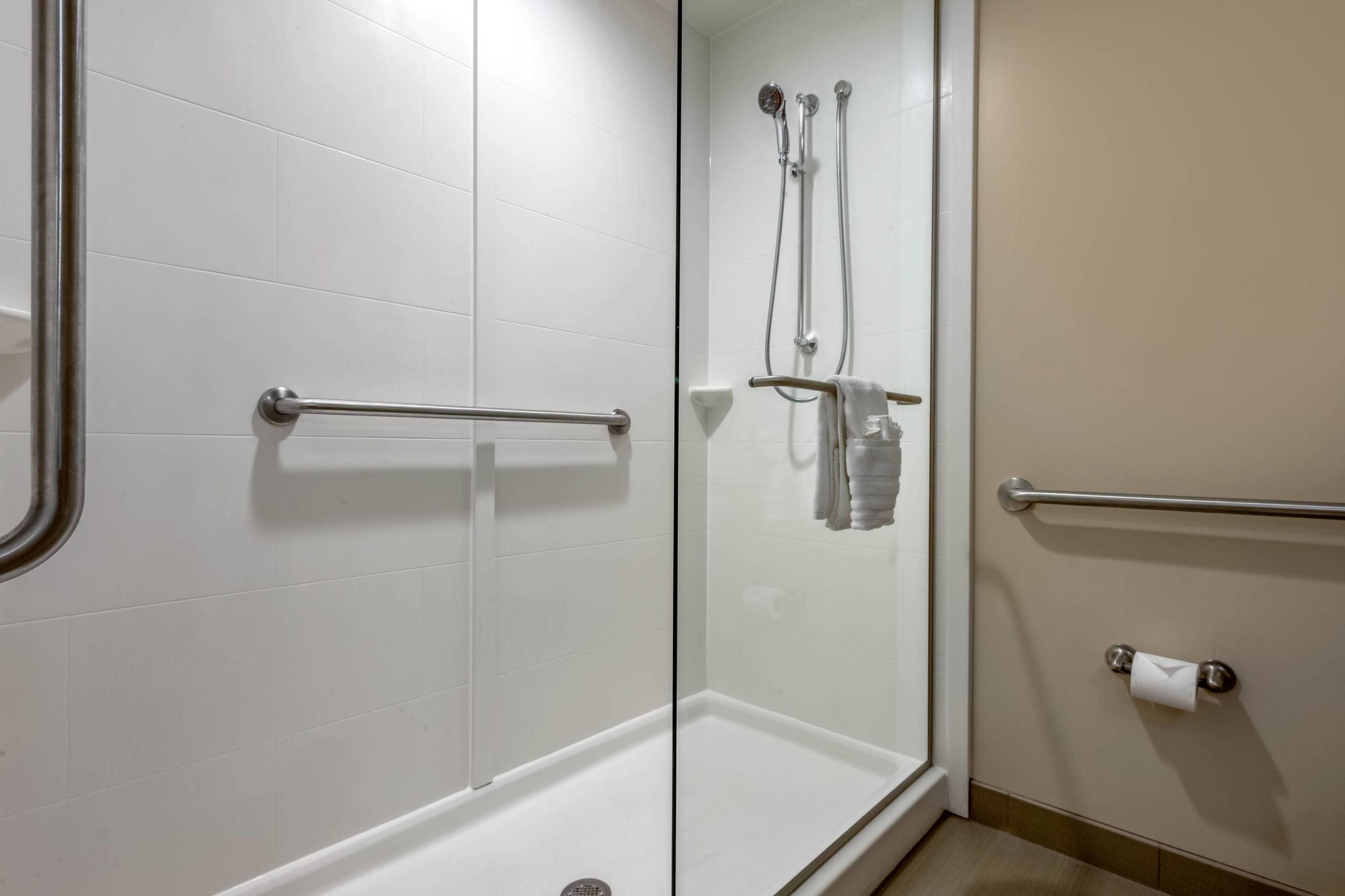 A modern bathroom with white tiles, a glass shower enclosure, a handrail, a towel rack, and a toilet paper holder.