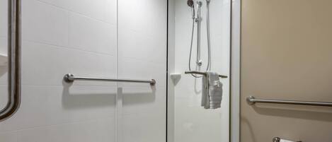A modern bathroom with white tiles, a glass shower enclosure, a handrail, a towel rack, and a toilet paper holder.