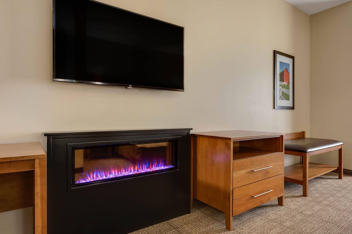 A room with a flat-screen TV, a fireplace with a visible fire, a wooden desk, a chair, and a framed picture on the wall.