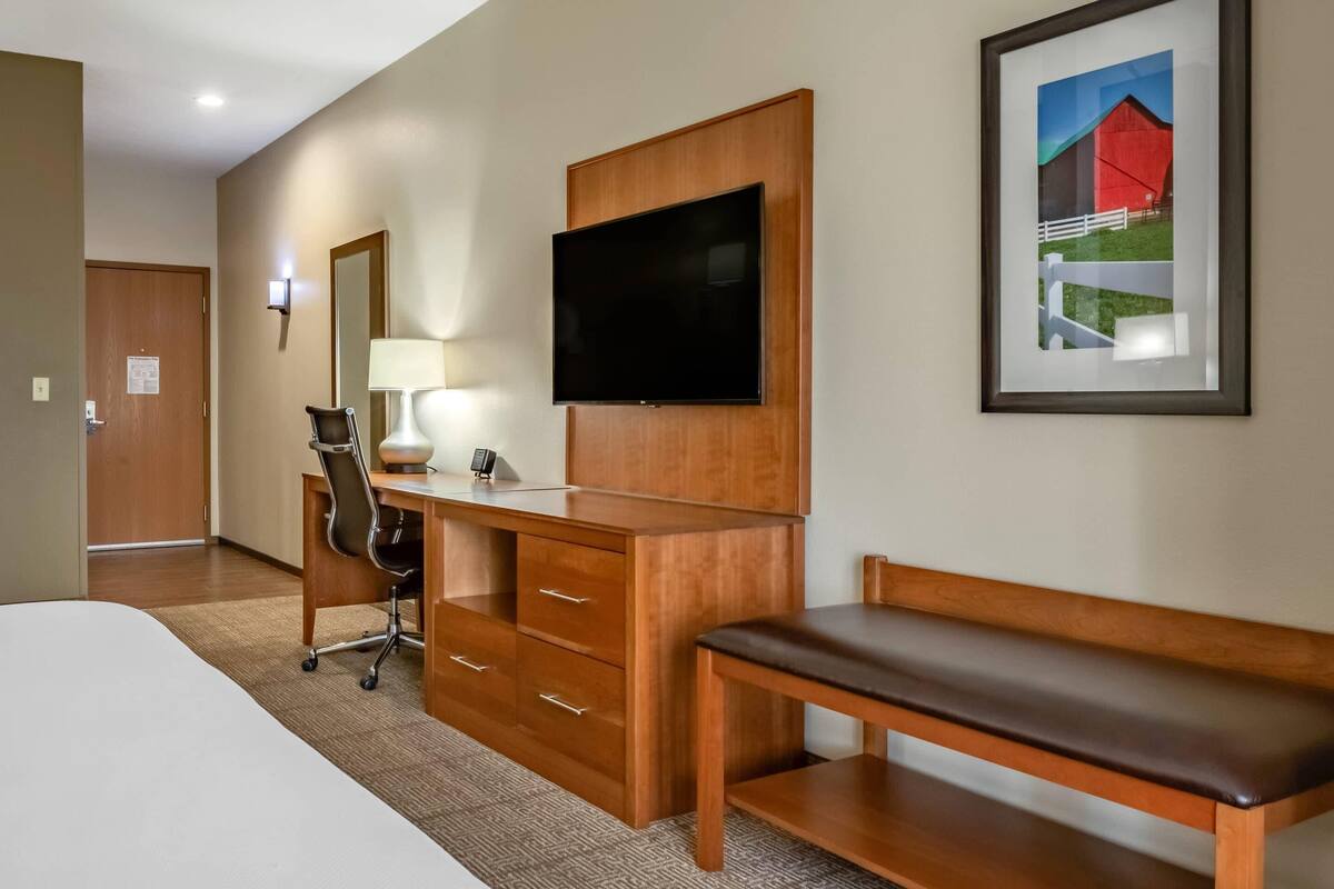 A hotel room with a bed, wooden desk, chair, TV, bench, and a framed picture on the wall.