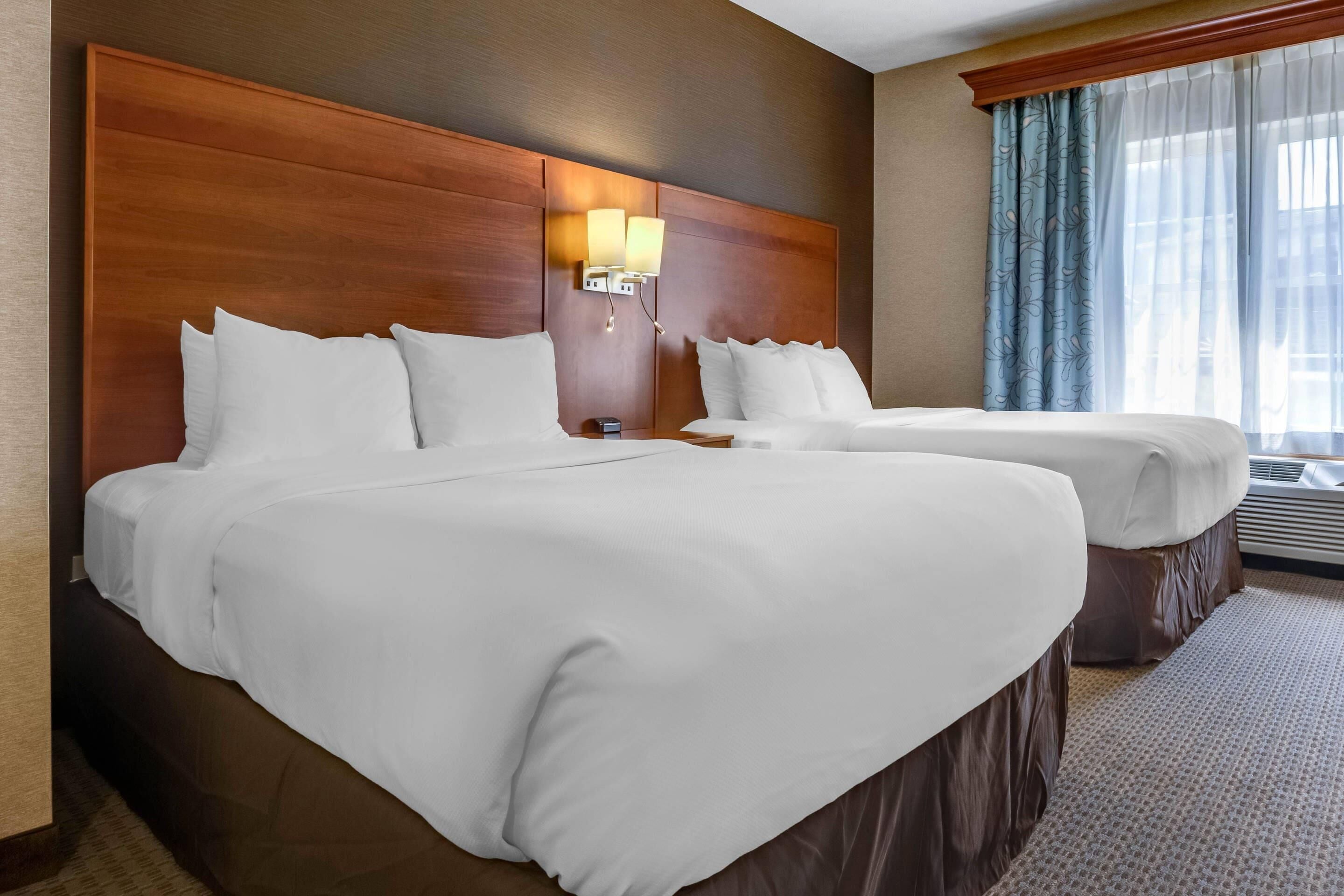 A hotel room with two beds, a wooden headboard, a nightstand, a lamp, and a window with curtains.
