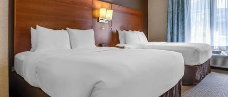 A hotel room with two beds, a wooden headboard, a nightstand, a lamp, and a window with curtains.