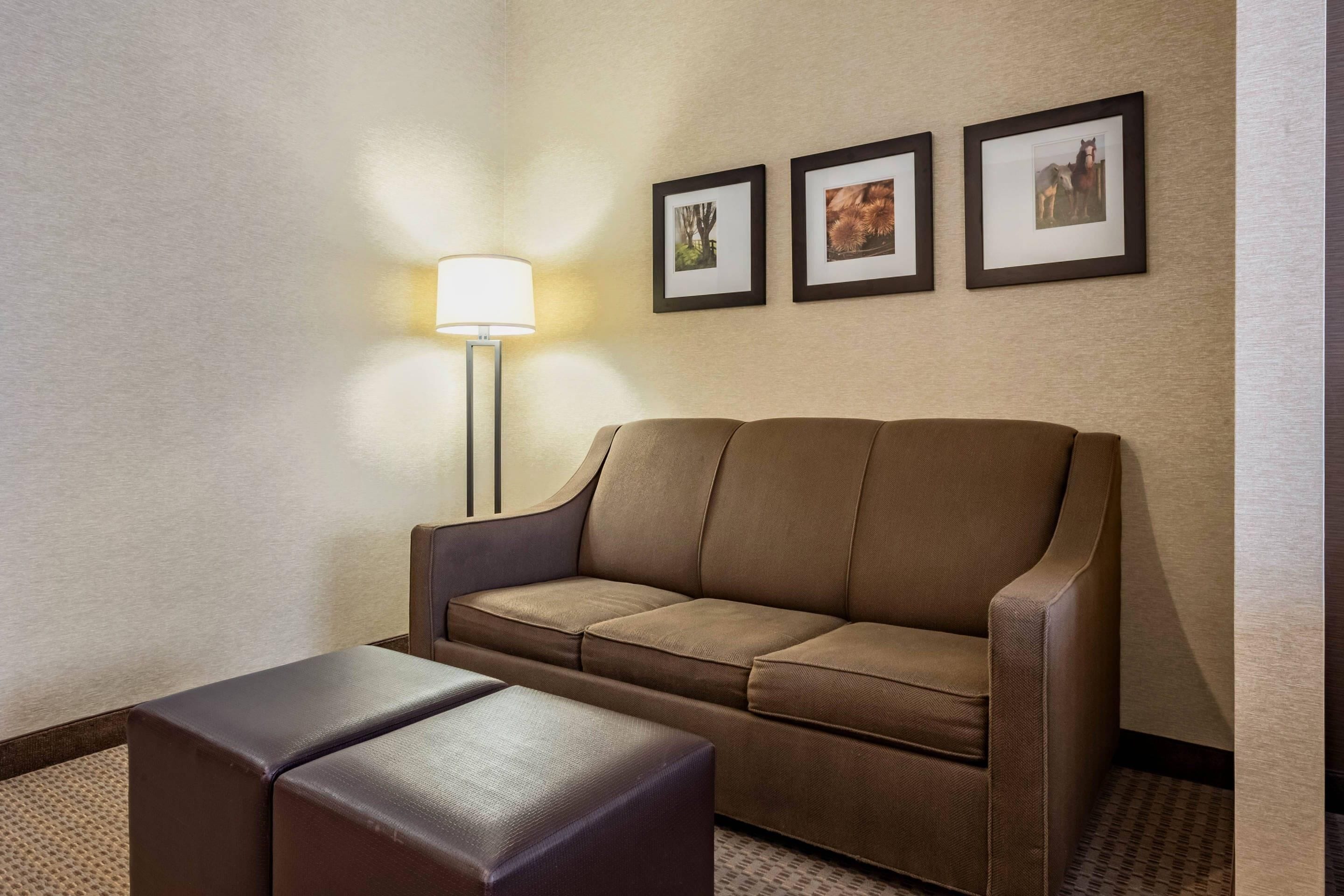 Ein Hotelzimmer mit braunem Sofa, einem kleinen Hocker, einer Stehlampe und drei gerahmten Bildern an der Wand.