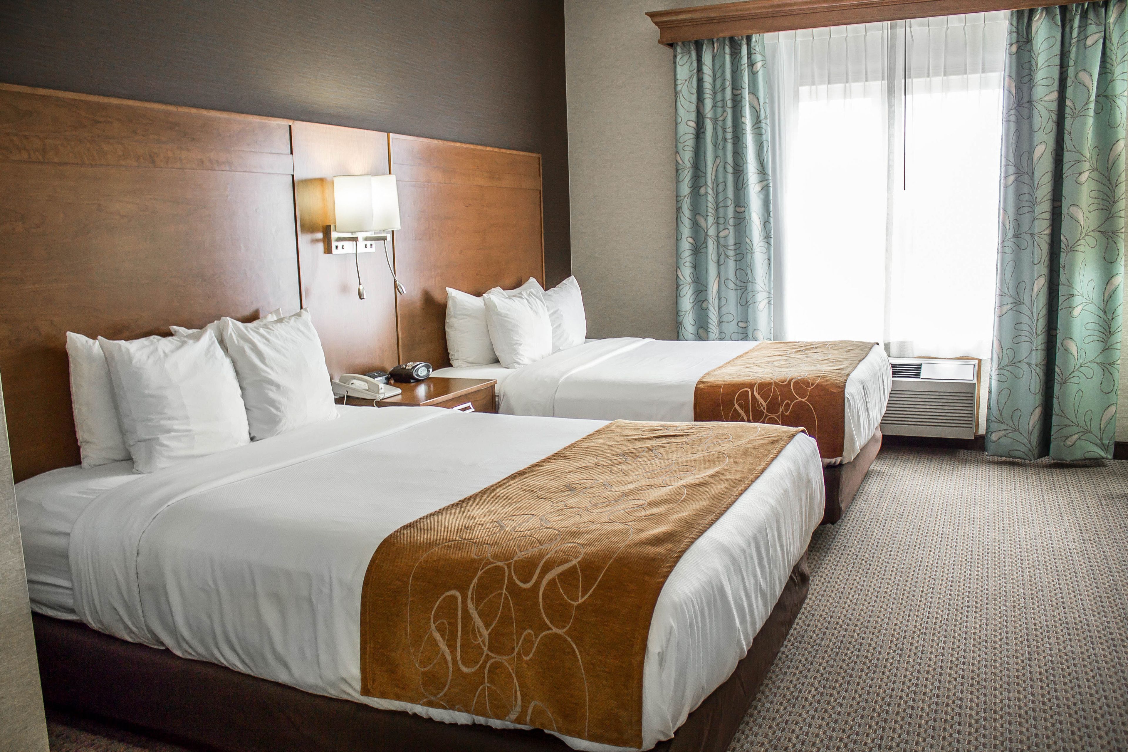 A hotel room with two beds, a wooden headboard, a bedside table with a lamp, and a window with curtains.