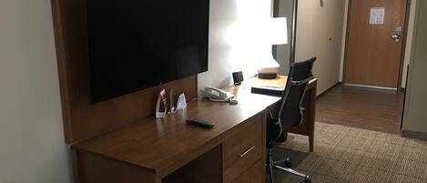 Habitación de hotel con un escritorio de madera, un televisor de pantalla plana, una lámpara, una silla y una puerta con un cartel.