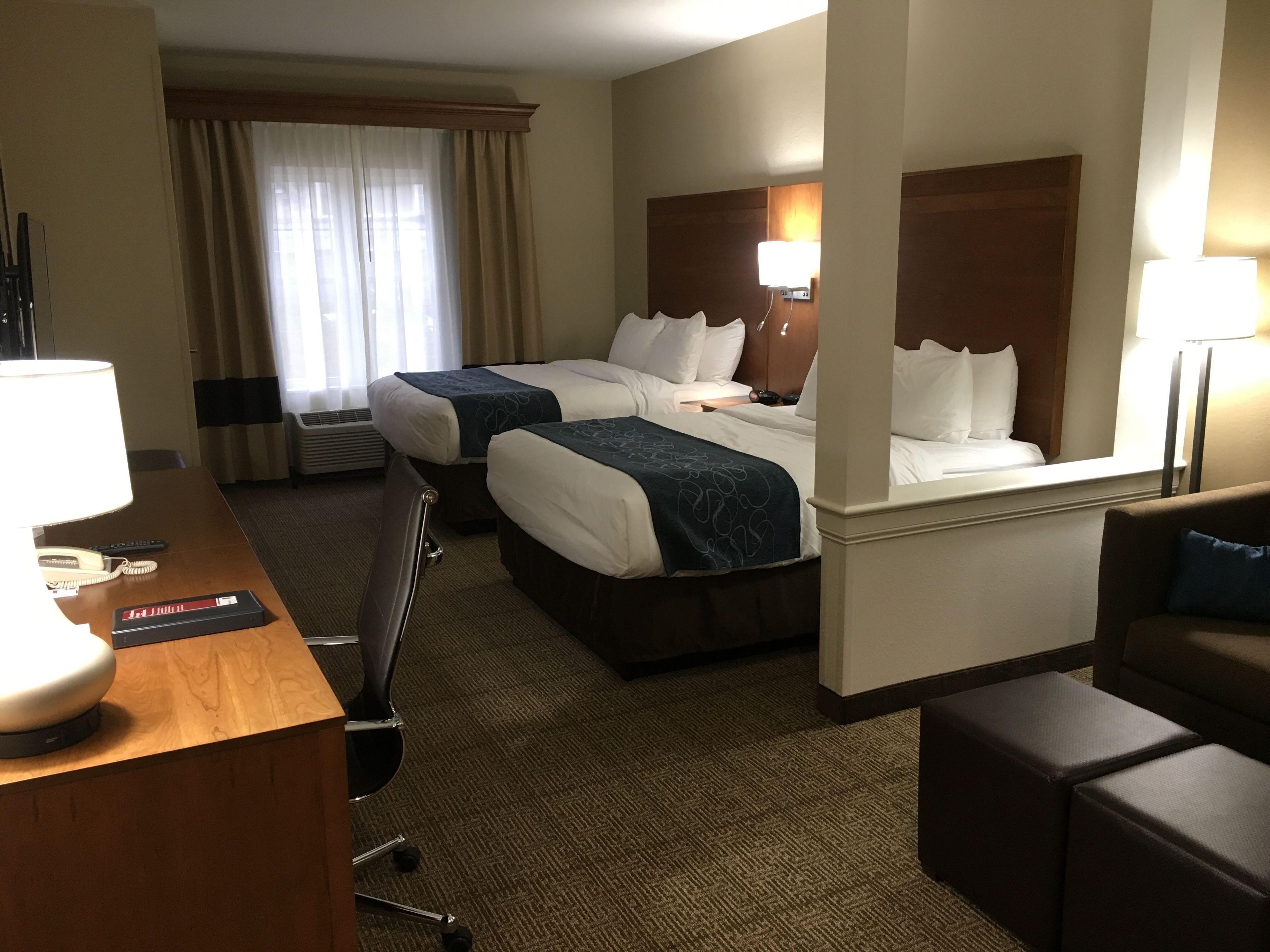 A hotel room with two beds, a desk with a lamp, a chair, a sofa, and a window with curtains.