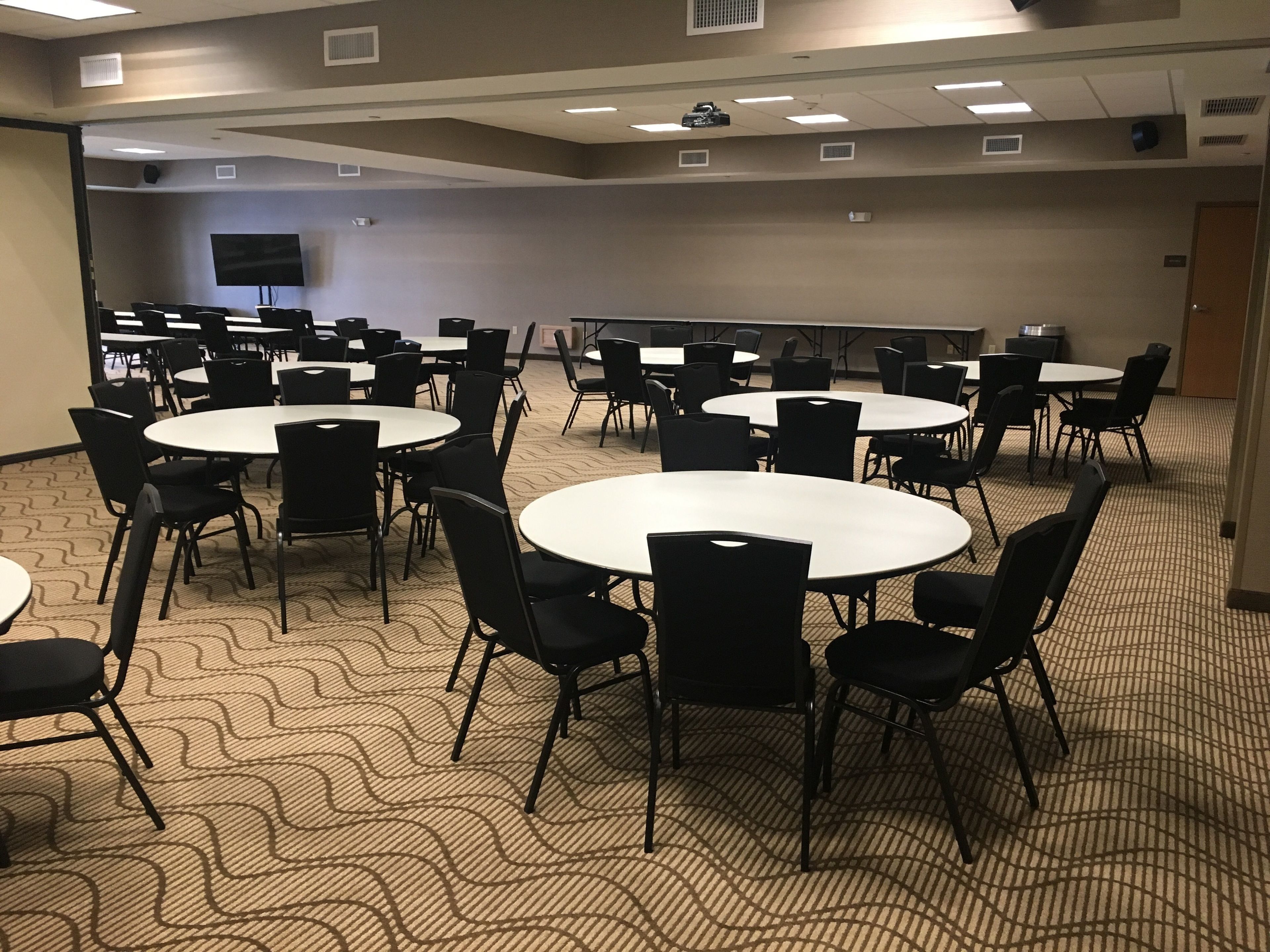 Un salón de conferencias con varias mesas y sillas, un proyector y una pantalla de televisión.