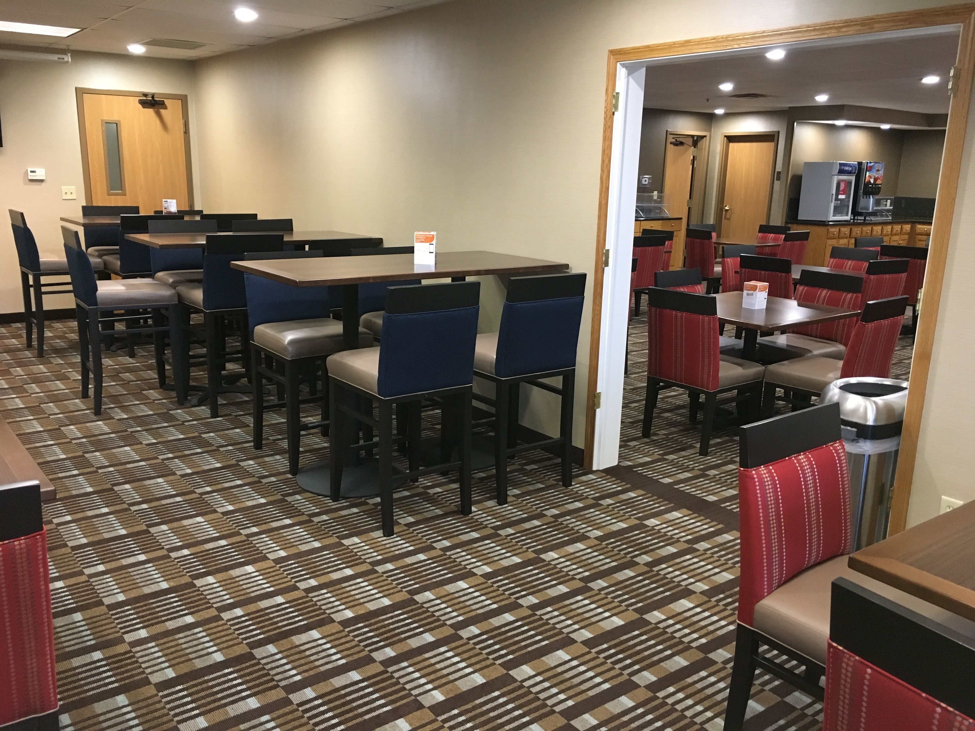 A hotel dining area with multiple tables and chairs, a patterned carpet, and a kitchen area visible through an open doorway.