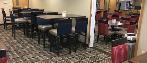 A hotel dining area with multiple tables and chairs, a patterned carpet, and a kitchen area visible through an open doorway.