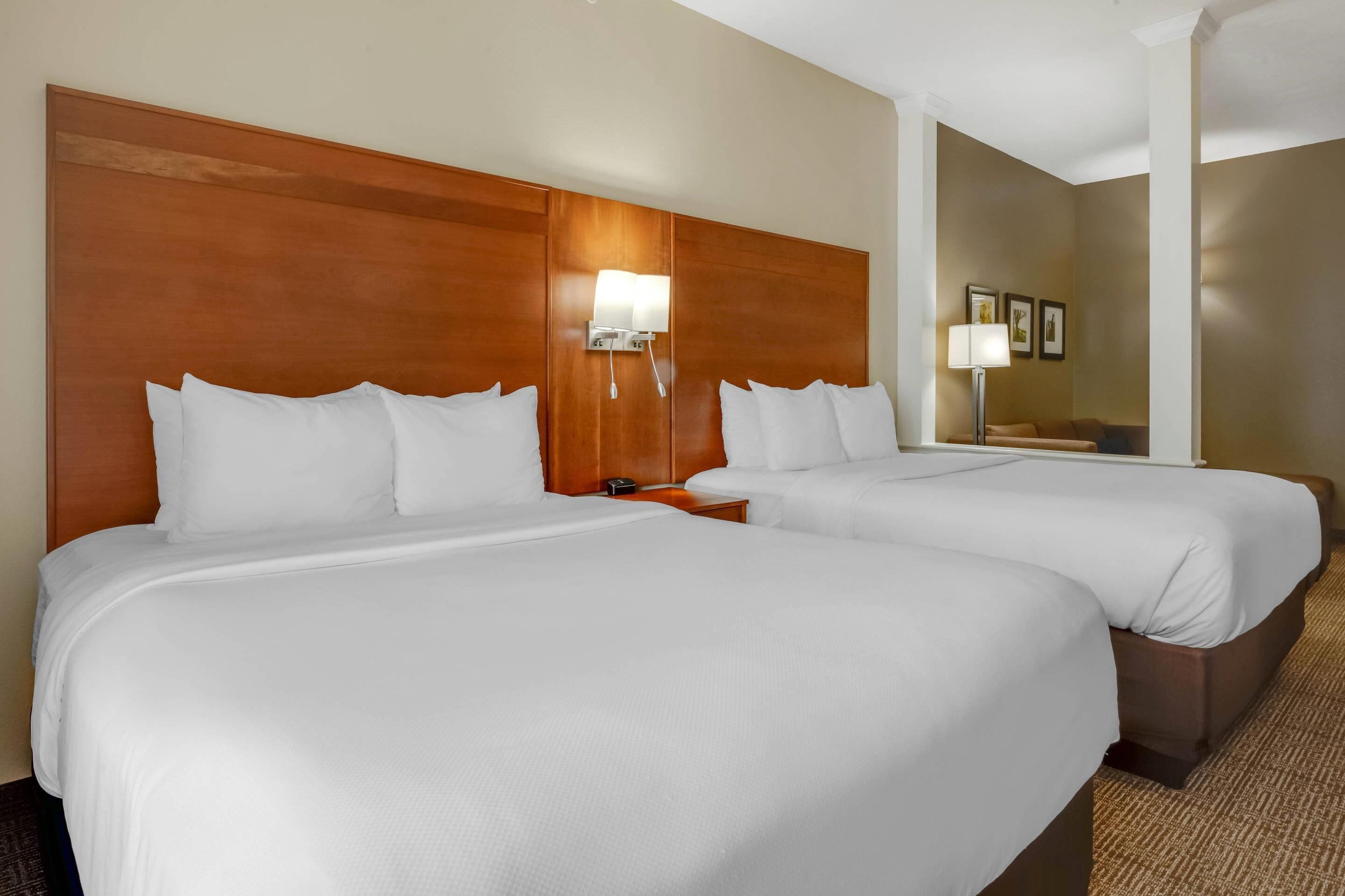 Habitación de hotel con dos camas, cabecero de madera, mesita de noche con lámpara y aplique de pared.
