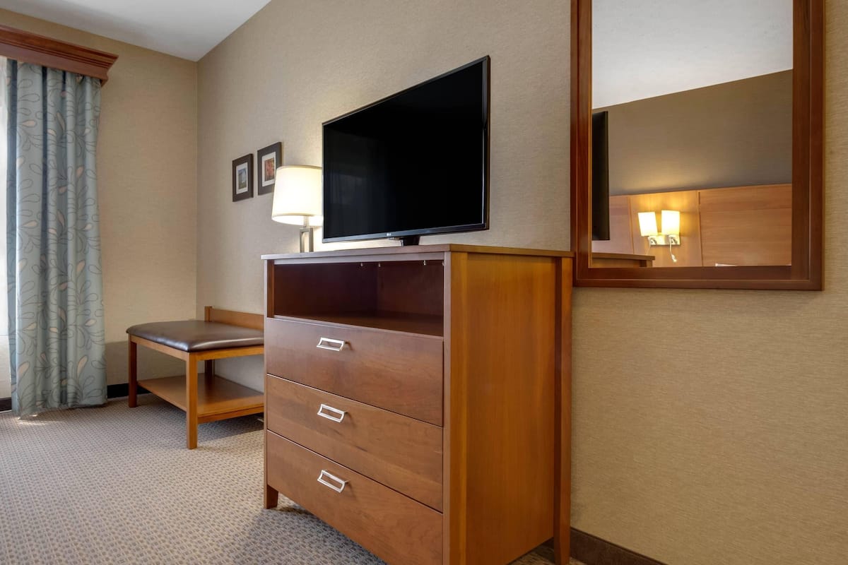 A hotel room with a wooden desk, a television on a stand, a mirror, and a lamp.