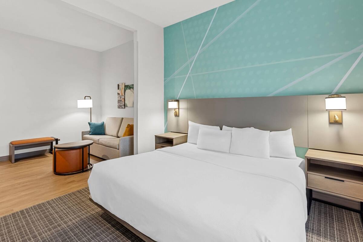 A hotel room with a large bed, two bedside tables, a bench, a sofa, and a wall with a geometric pattern.