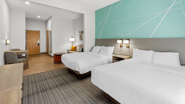 A hotel room with two beds, a desk, a chair, a small table, and a wall-mounted TV.
