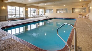 Una piscina interior con agua cristalina, paredes de azulejos y una sala iluminada con ventanas y sillones de descanso.