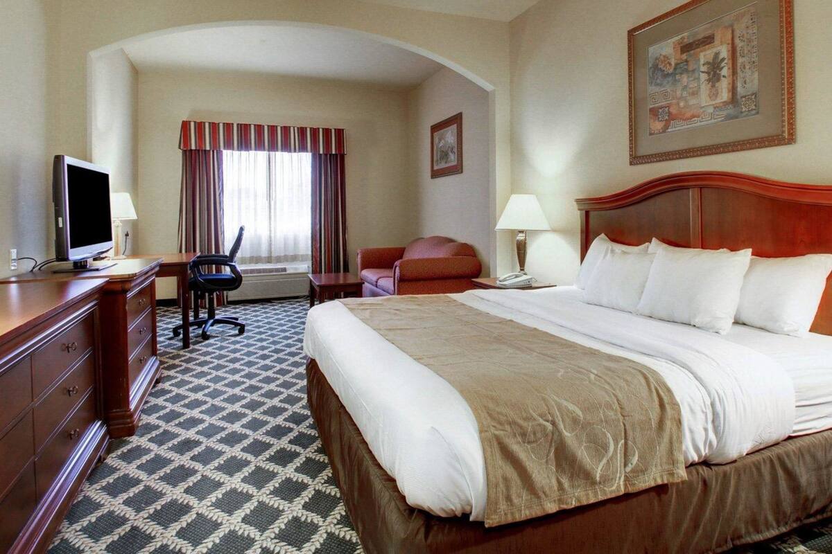 A hotel room with a large bed, a desk with a computer, a chair, a sofa, and a TV.