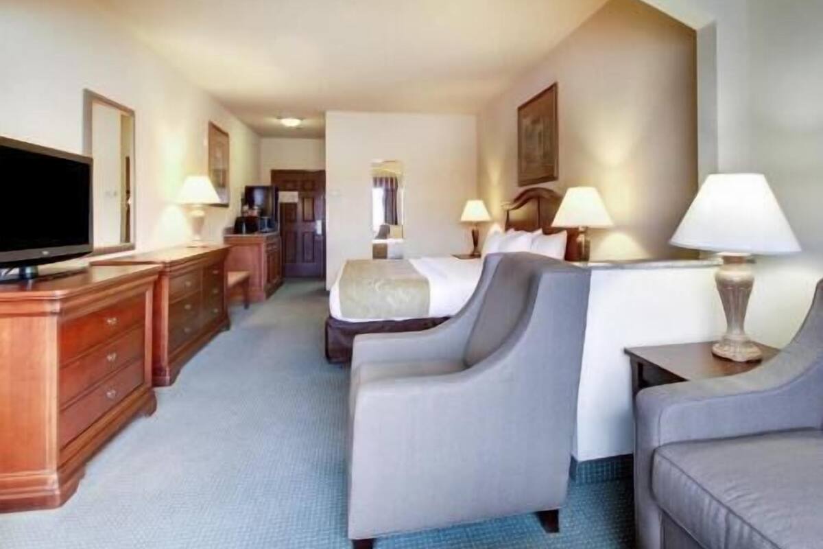 A hotel room with a television, a desk, two chairs, a bed, and a nightstand with lamps.