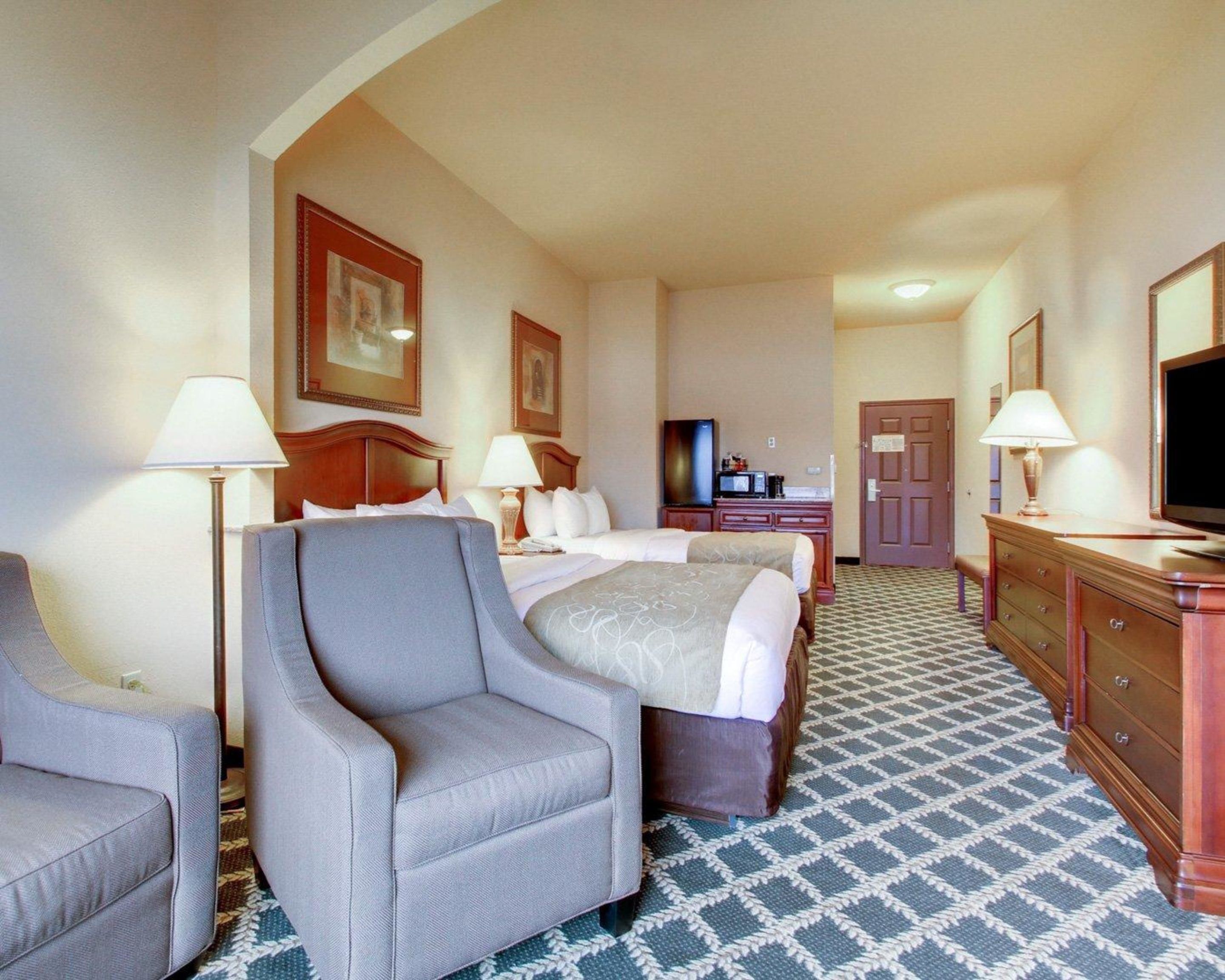 A hotel room with a bed, two armchairs, a dresser, a television, and a door.
