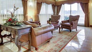 Amplia sala de estar con un sofá de cuero, dos sillones estampados, una mesa de centro de madera y un arreglo decorativo de plantas. Grandes ventanas con cortinas ofrecen luz natural y vistas.