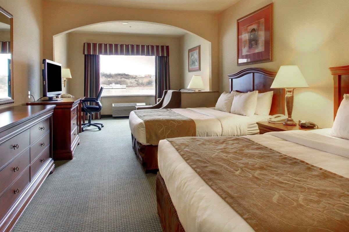 A hotel room with two beds, a desk with a computer, a chair, and a view of the outdoors.