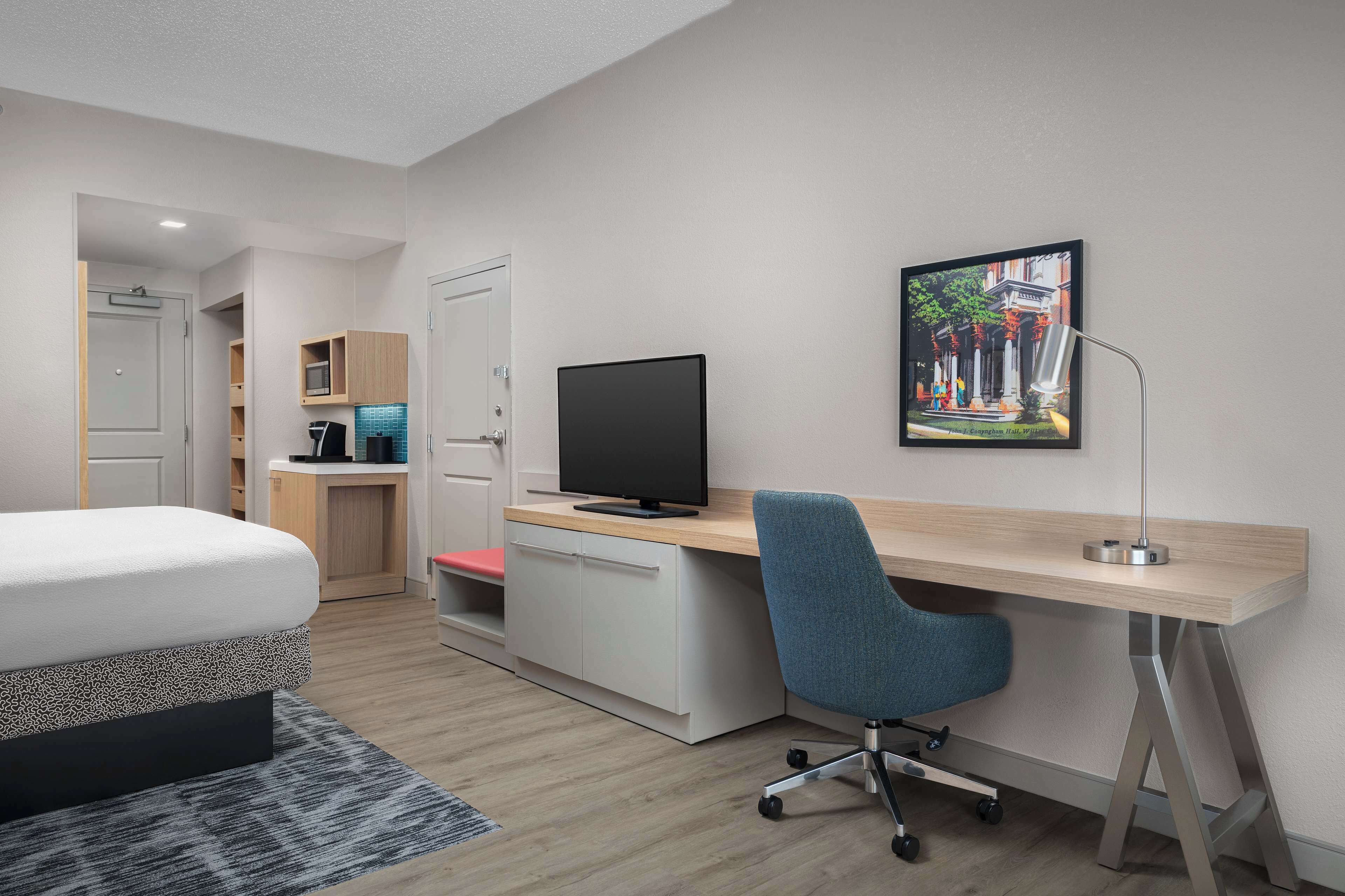 A modern hotel room with a bed, a desk with a computer, a TV, and a picture of a cityscape on the wall.