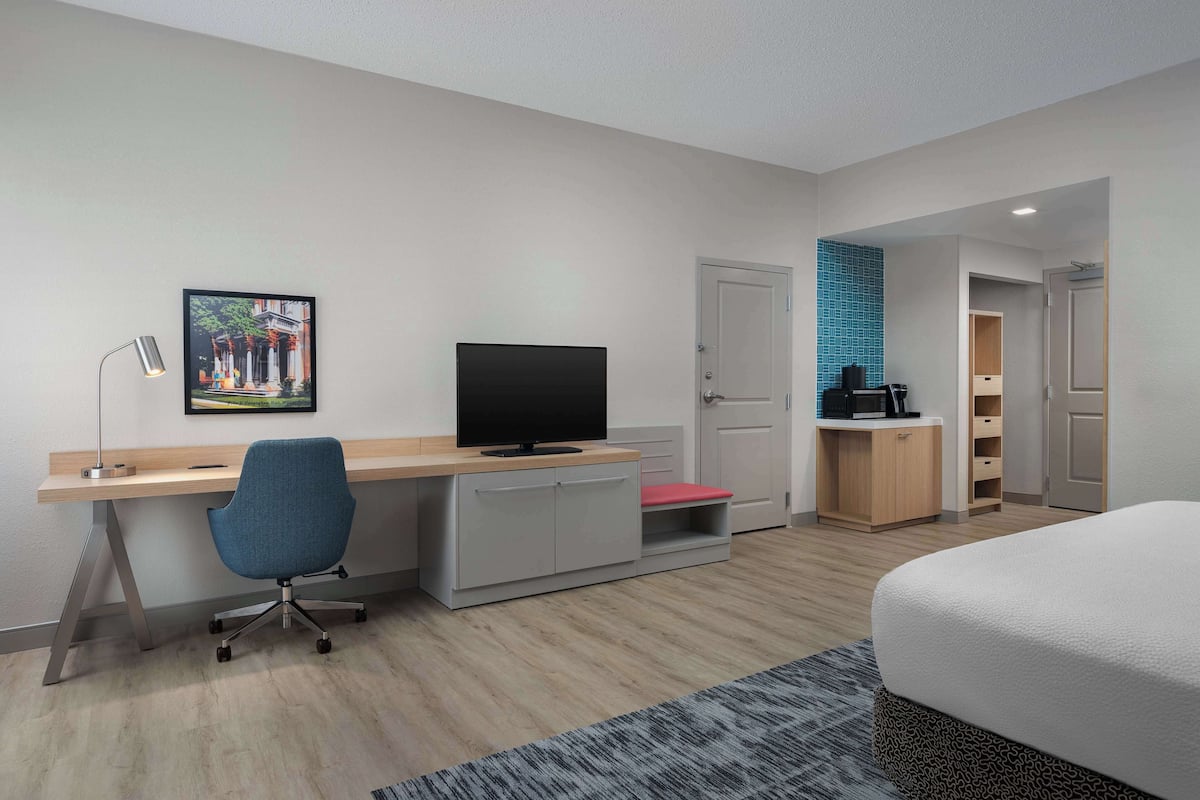 A modern hotel room with a wooden desk, a blue office chair, a flat-screen TV, a bed with a patterned bedspread, and a small red bench.