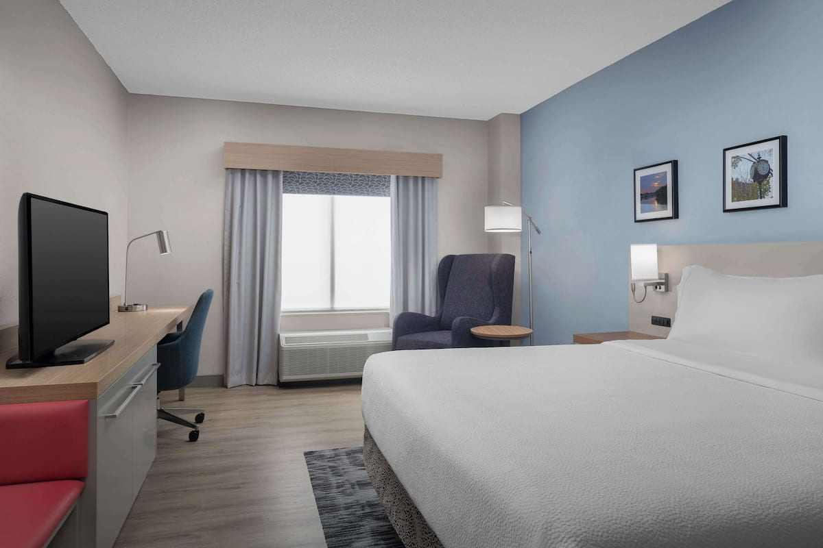 A modern hotel room with a large bed, a desk with a flat-screen TV, a comfortable armchair, and a small side table.