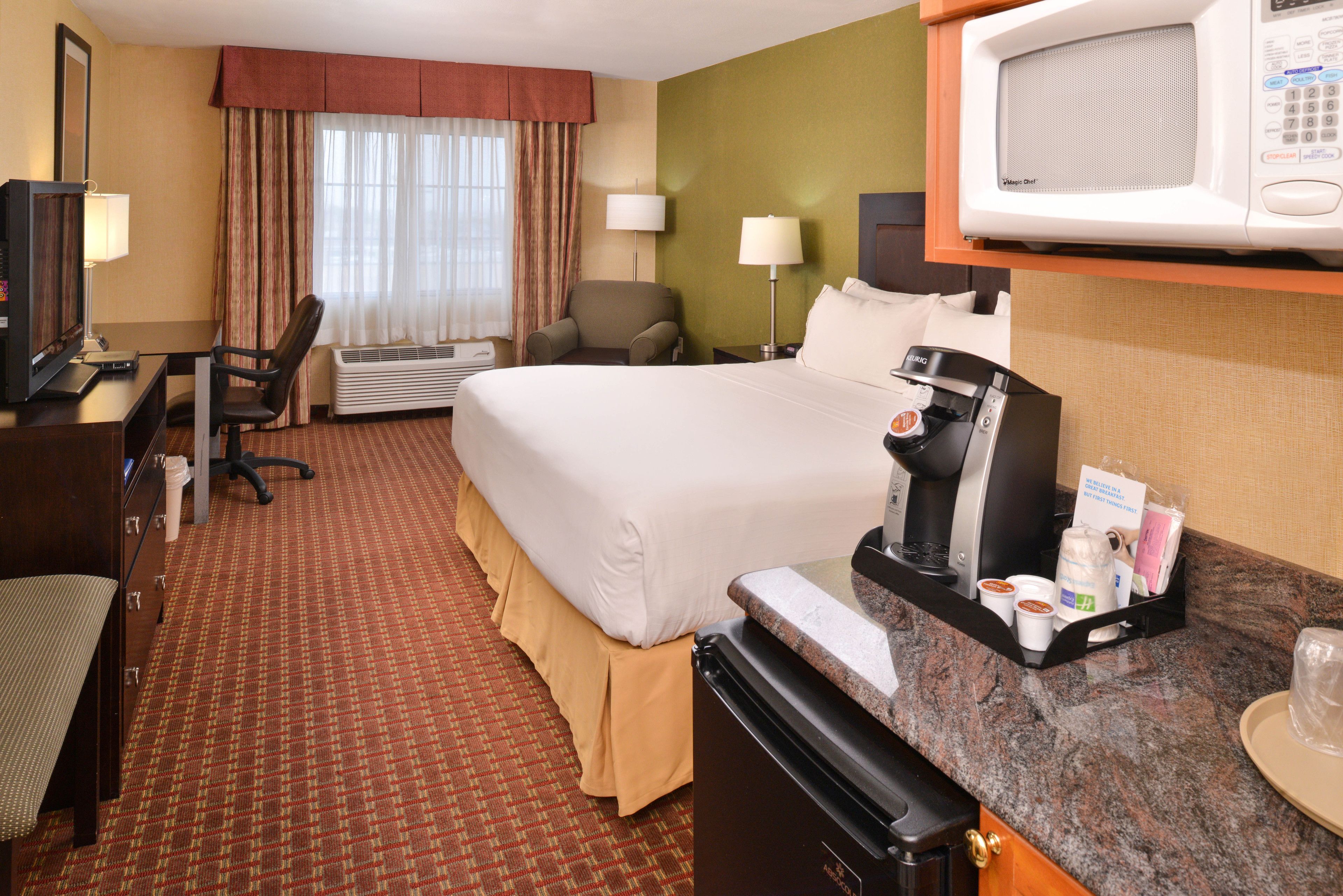 A hotel room with a bed, desk, chair, microwave, coffee maker, and a small kitchenette.