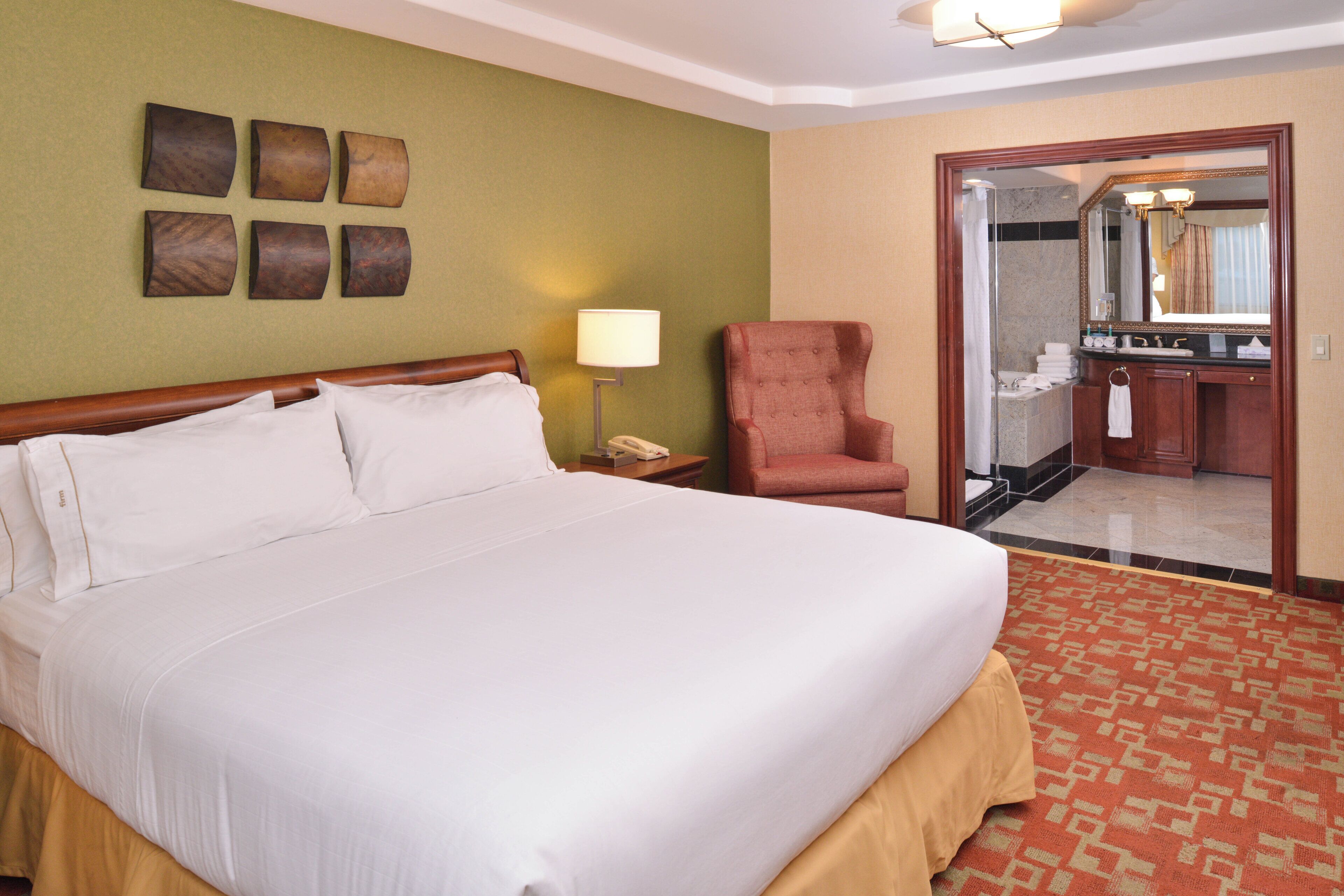 Ein Hotelzimmer mit einem großen Bett, einem Sessel, einem Schreibtisch mit Lampe und einem durchgehenden Türspalte sichtbaren Badezimmer.