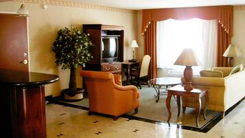 Habitación de hotel con una mesa redonda, un sofá, un sillón, una planta en maceta, un televisor y una lámpara.