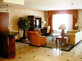 Habitación de hotel con una mesa redonda, un sofá, un sillón, una planta en maceta, un televisor y una lámpara.
