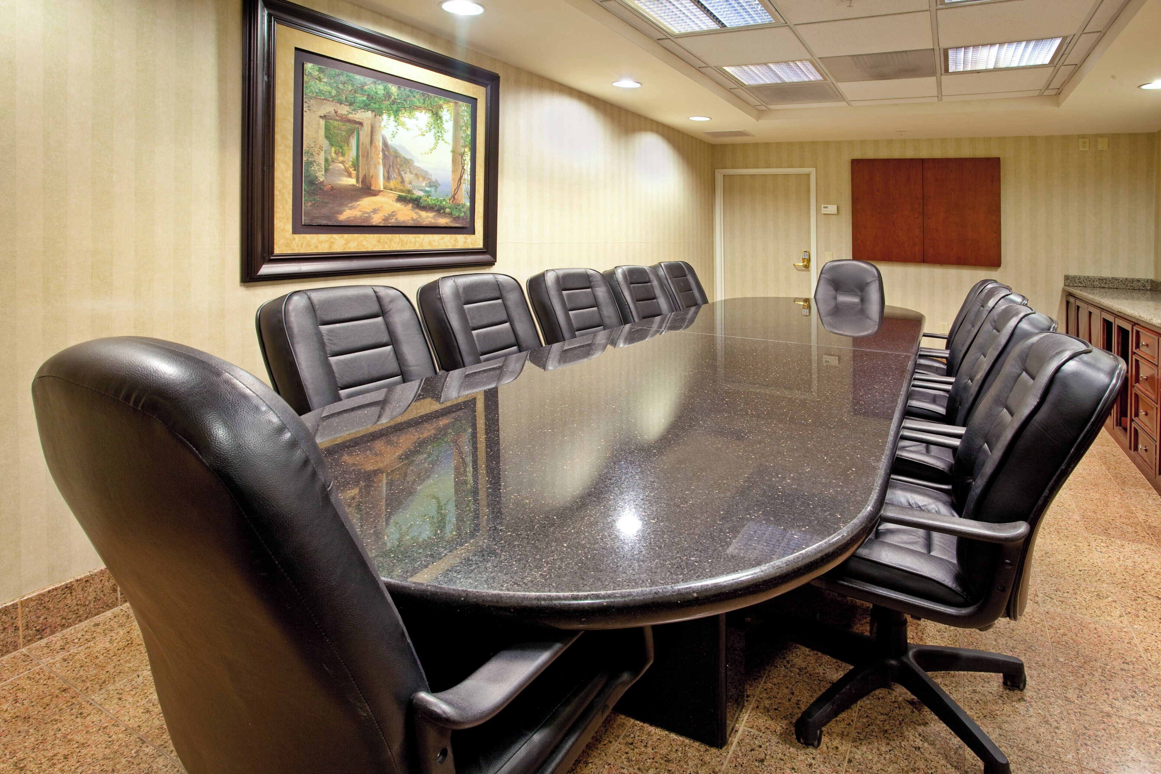 A conference room with a long table surrounded by leather chairs, a framed painting on the wall, and a door with a wooden panel.