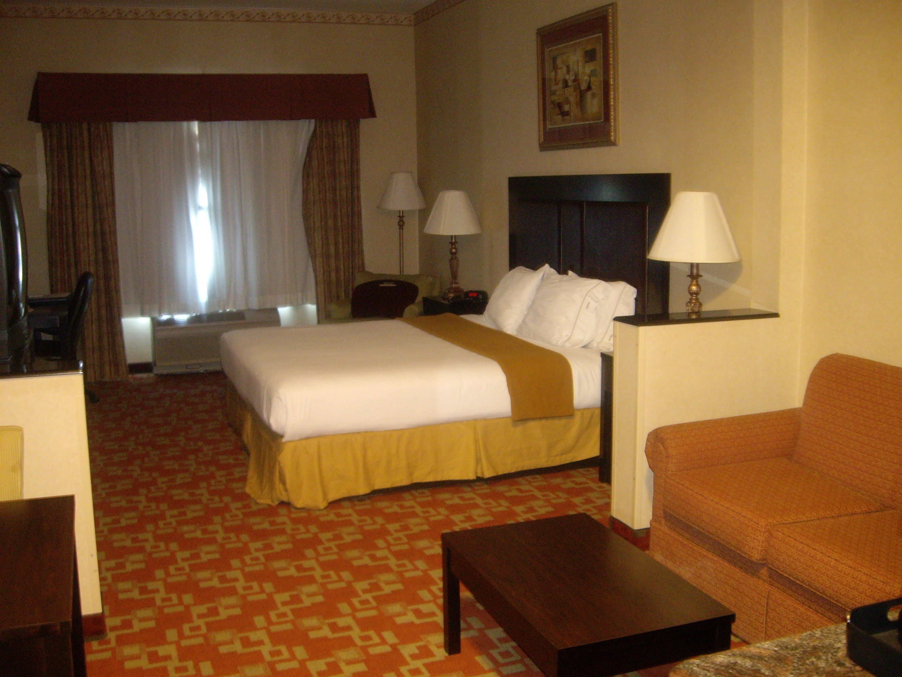 A hotel room with a bed, two bedside lamps, a sofa, a coffee table, a chair, and a large window with curtains.