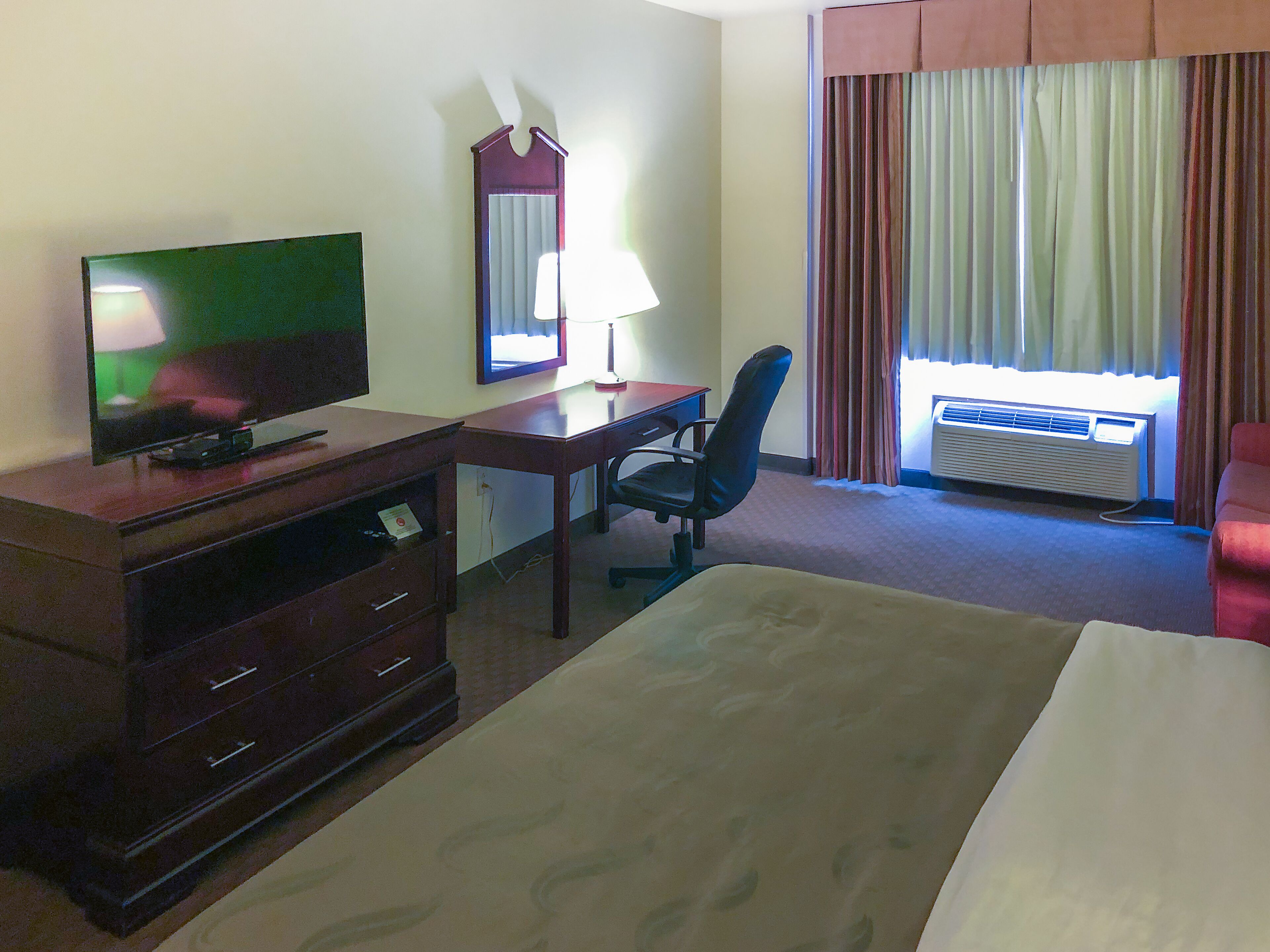Habitación de hotel con cama, escritorio con silla, televisor, espejo y aire acondicionado.