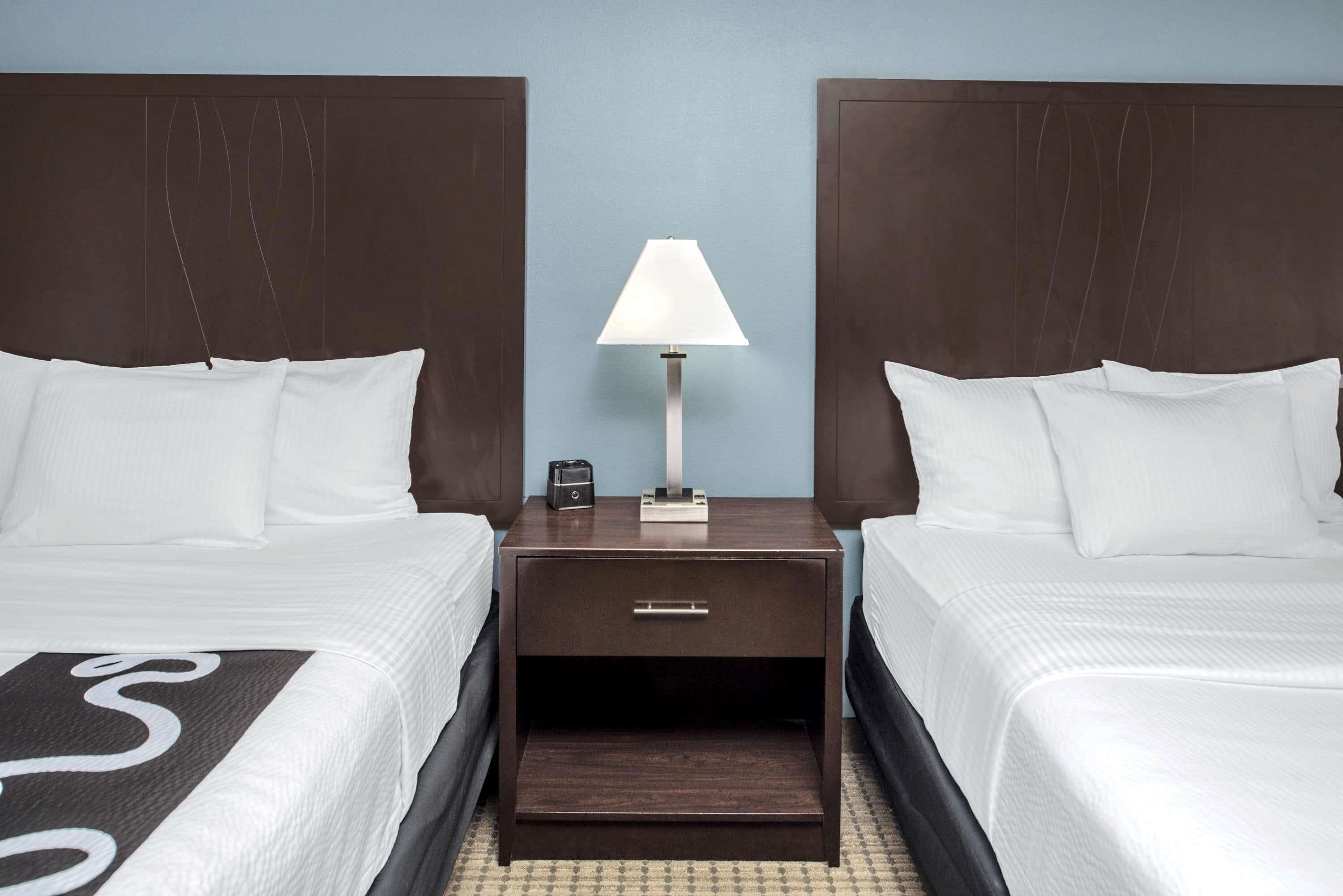 A hotel room with two beds, a nightstand with a lamp, and a small drawer.