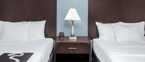 A hotel room with two beds, a nightstand with a lamp, and a small drawer.