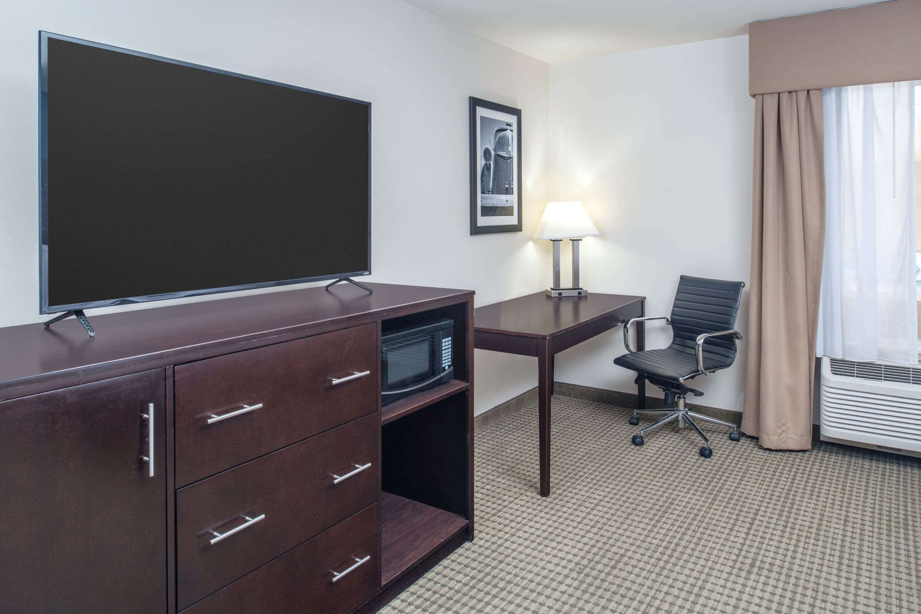 A hotel room with a flat-screen TV, a desk with a lamp, a chair, a microwave, and a large cabinet with drawers.