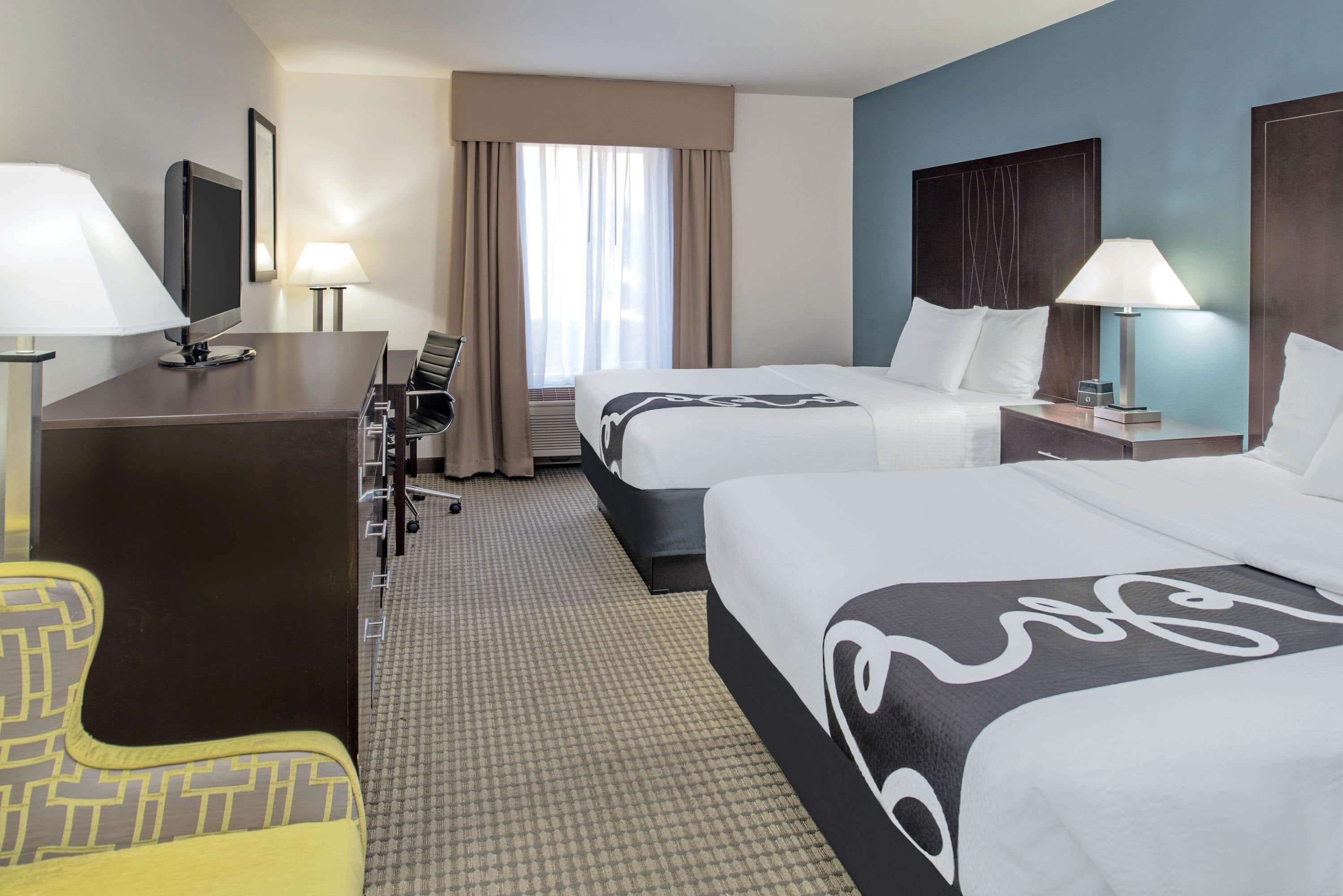Quarto de hotel com duas camas, uma escrivaninha, uma cadeira, uma televisão e uma janela com cortinas.