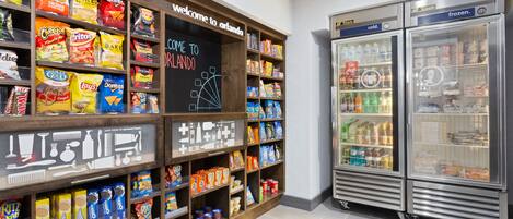Exposição de lanches e bebidas, com prateleiras repletas de diversos produtos e um refrigerador de duas portas.