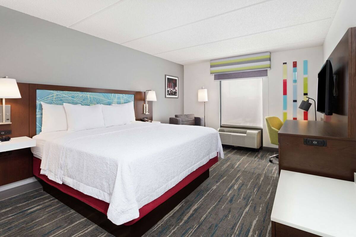 A hotel room with a large bed, a desk, a chair, a TV, and a window with blinds.