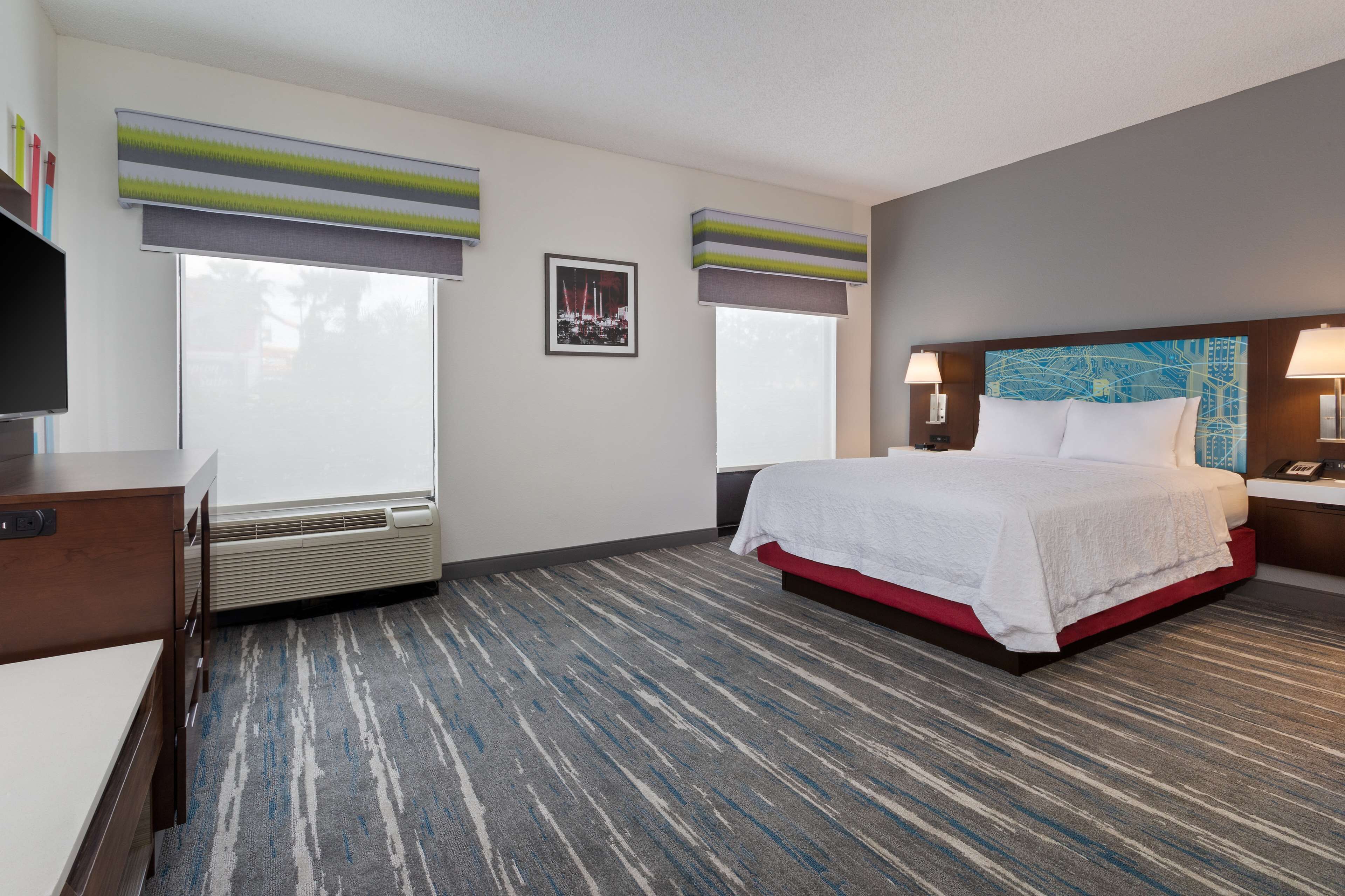 A hotel room with a bed, bedside tables, a desk, and a large window with blinds.
