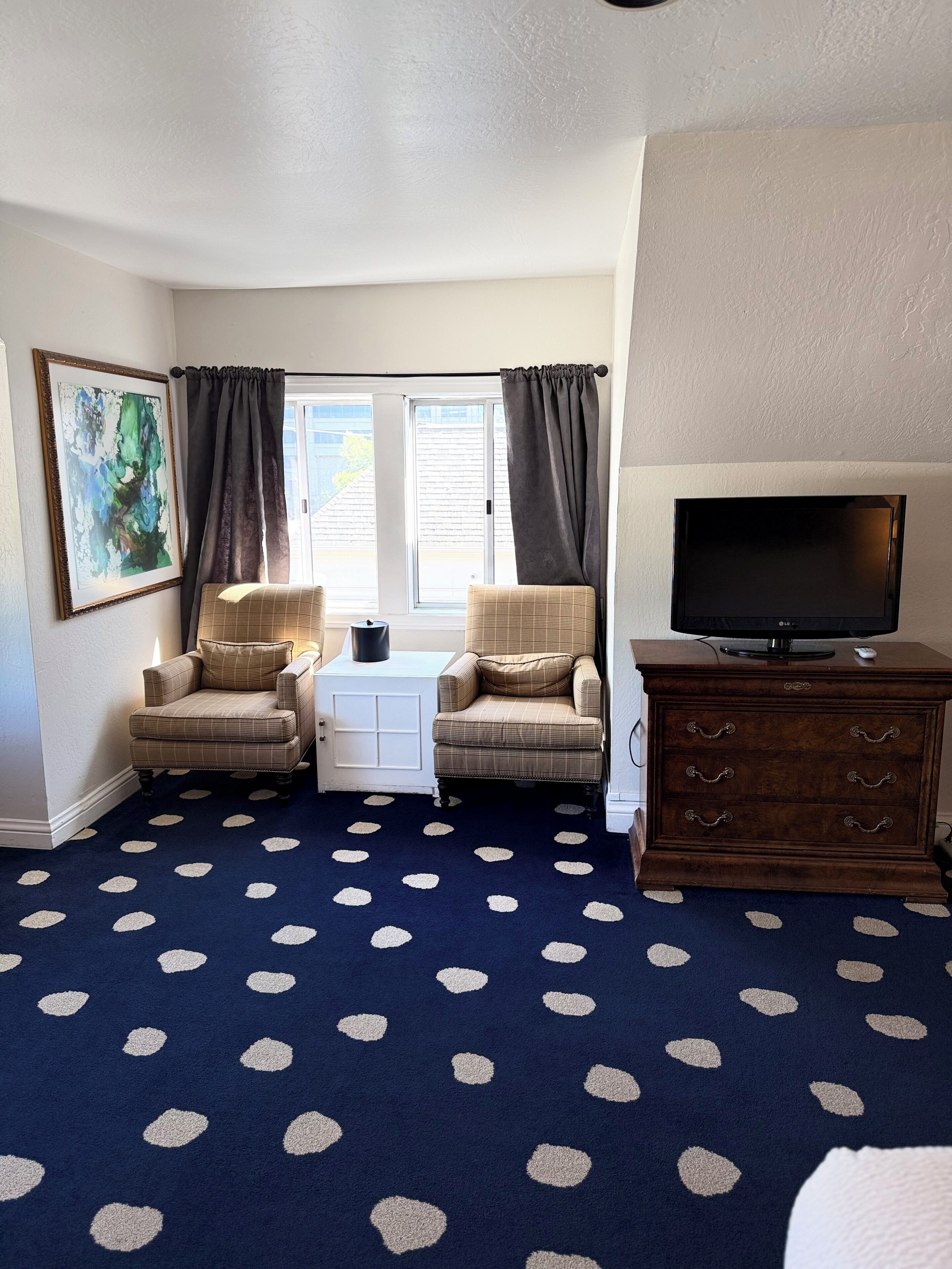 A room with a dark blue carpet with white polka dots, two armchairs, a small white table, a television on a wooden cabinet, and a window with curtains.