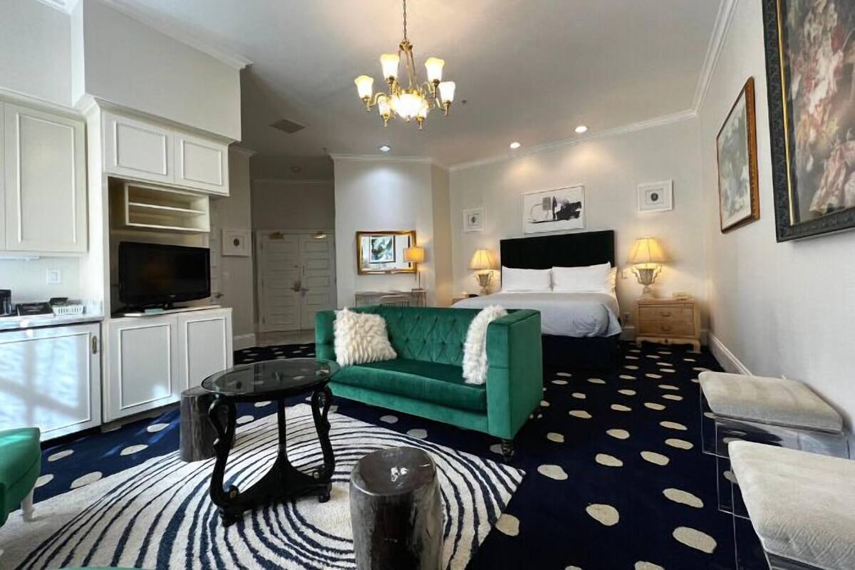 A hotel room with a green sofa, a round coffee table, a TV, a bed with two pillows, a nightstand with lamps, and a polka dot carpet.