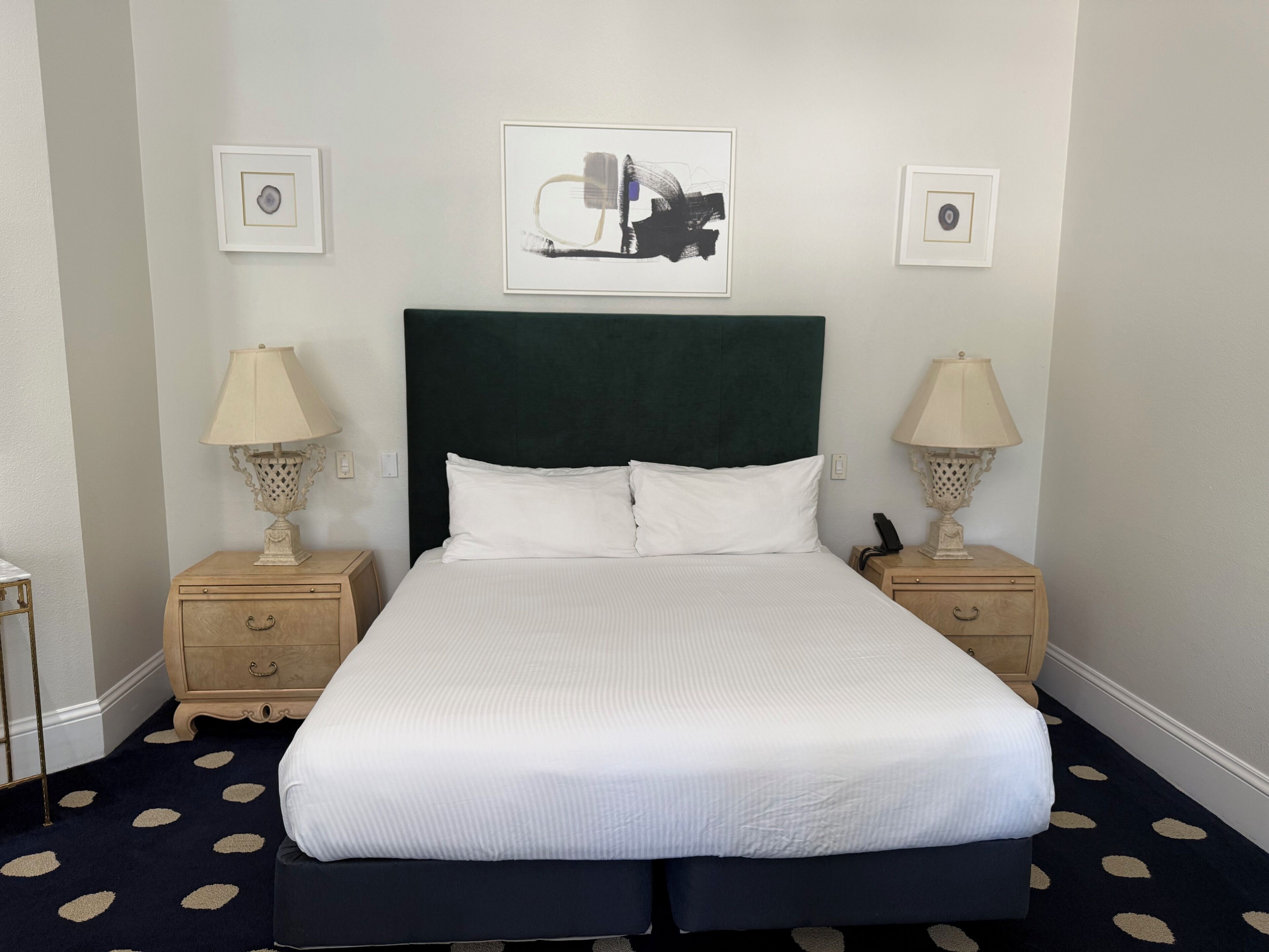 Habitación de hotel con una cama, dos mesitas de noche con lámparas, un cabecero y dos obras de arte enmarcadas en la pared.