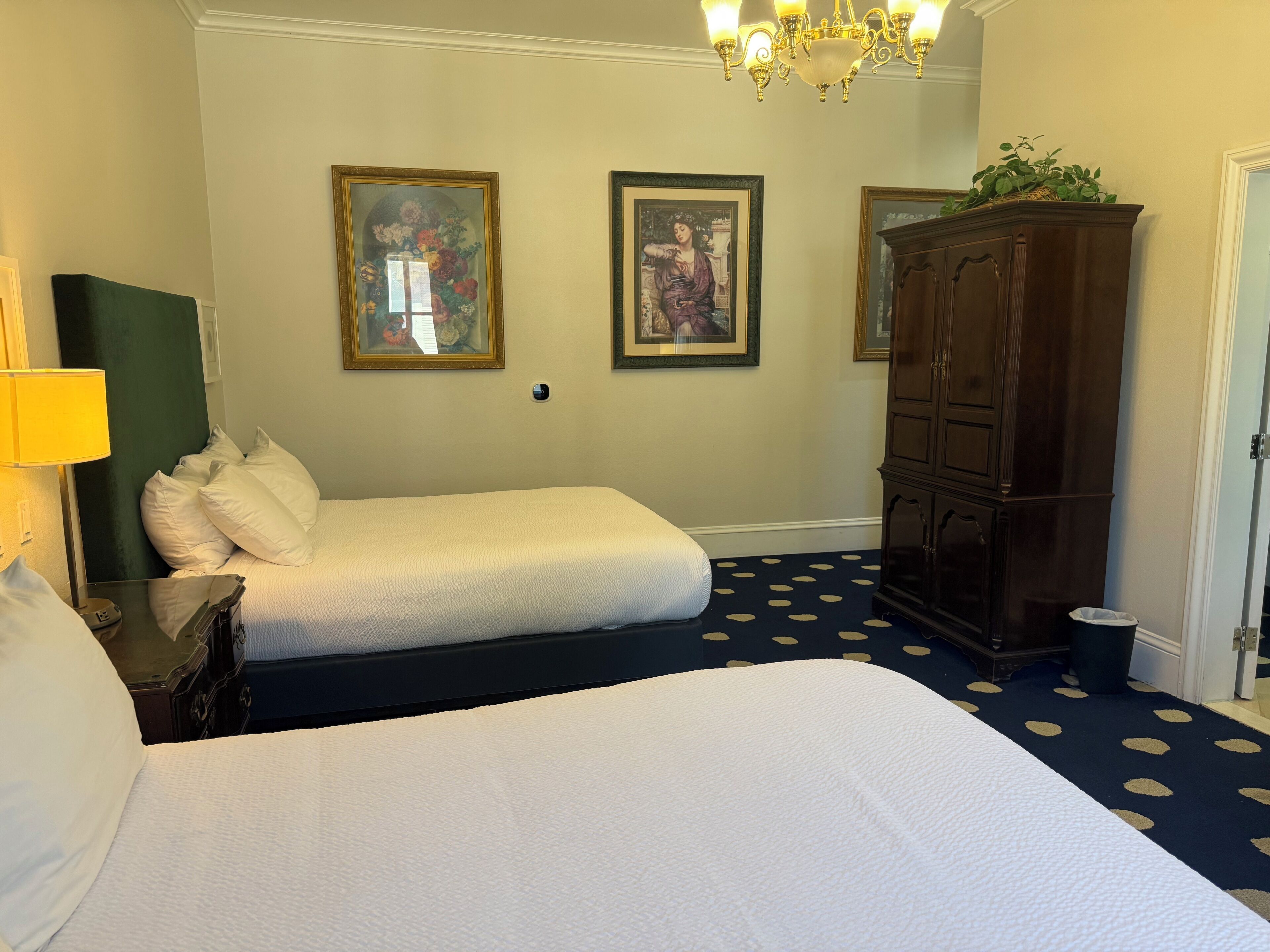 A hotel room with two beds, a chandelier, a cabinet, and two framed pictures on the wall.