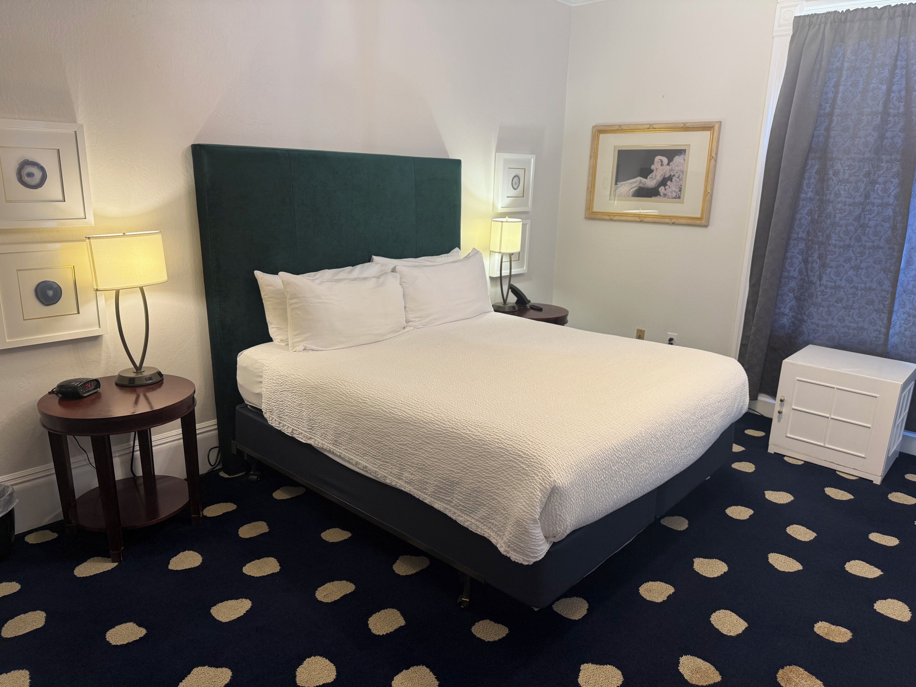 A hotel room with a bed, bedside lamps, a nightstand, a small cabinet, and a patterned carpet.